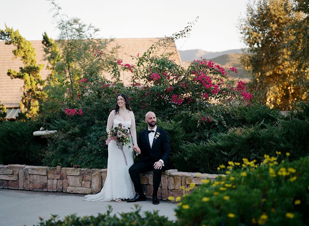 A bride and groom outdoors, with the bride in a white wedding dress holding a bouquet, standing next to a seated groom in a tuxedo, surrounded by greenery and pink flowering bushes.