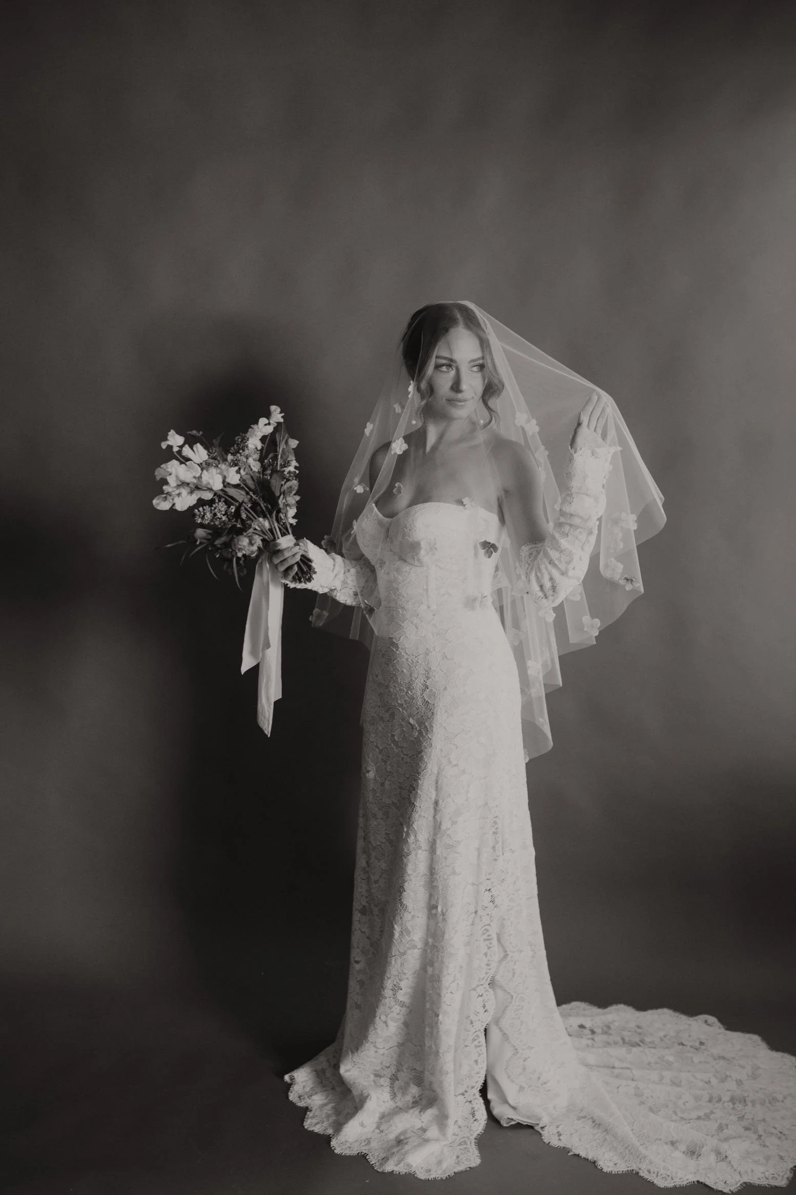 Black and white photo of a bride in a lace wedding gown holding a bouquet of flowers and lifting her veil.