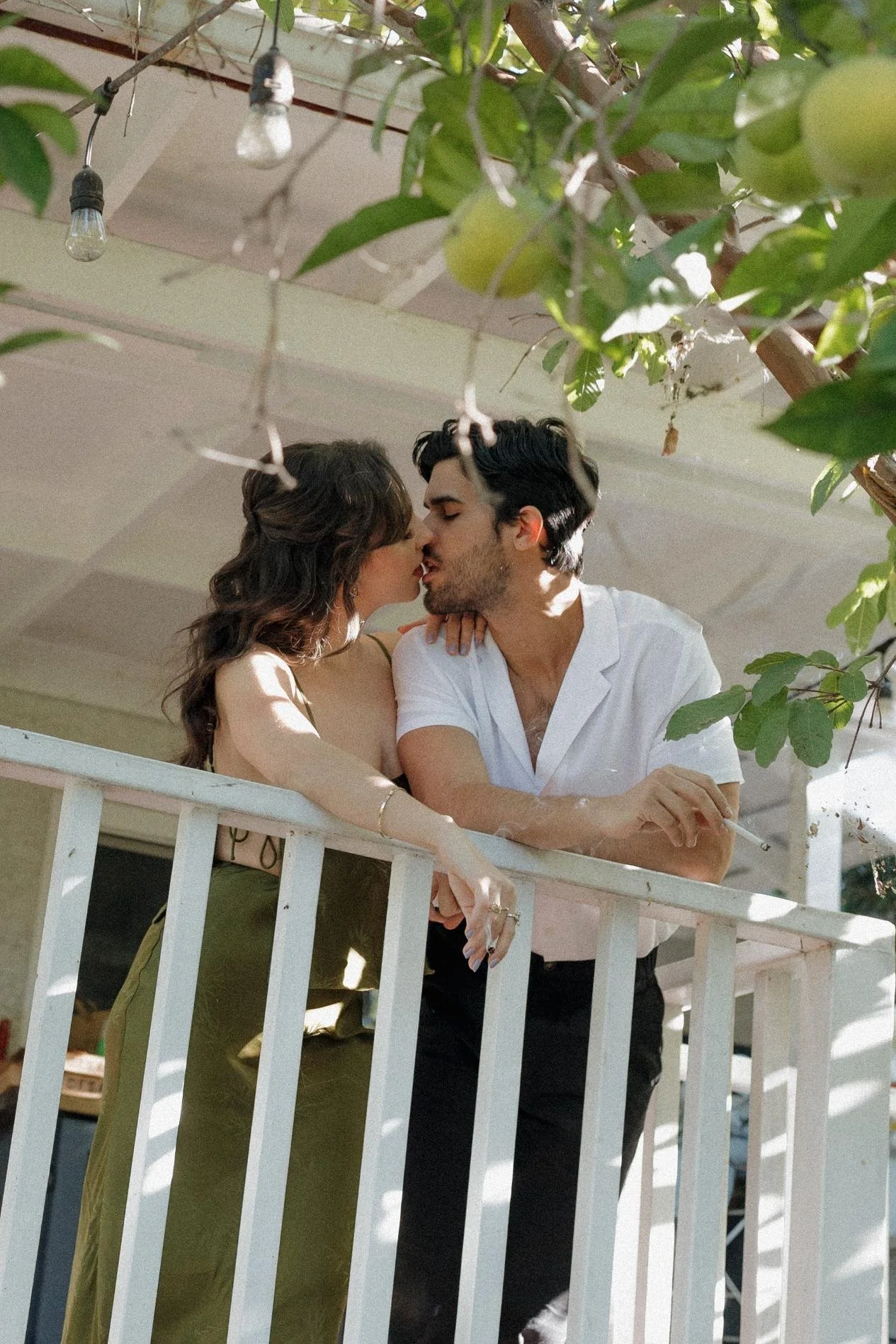 A young couple sharing a kiss on a balcony, surrounded by green leaves and string lights, with sunlight casting shadows.