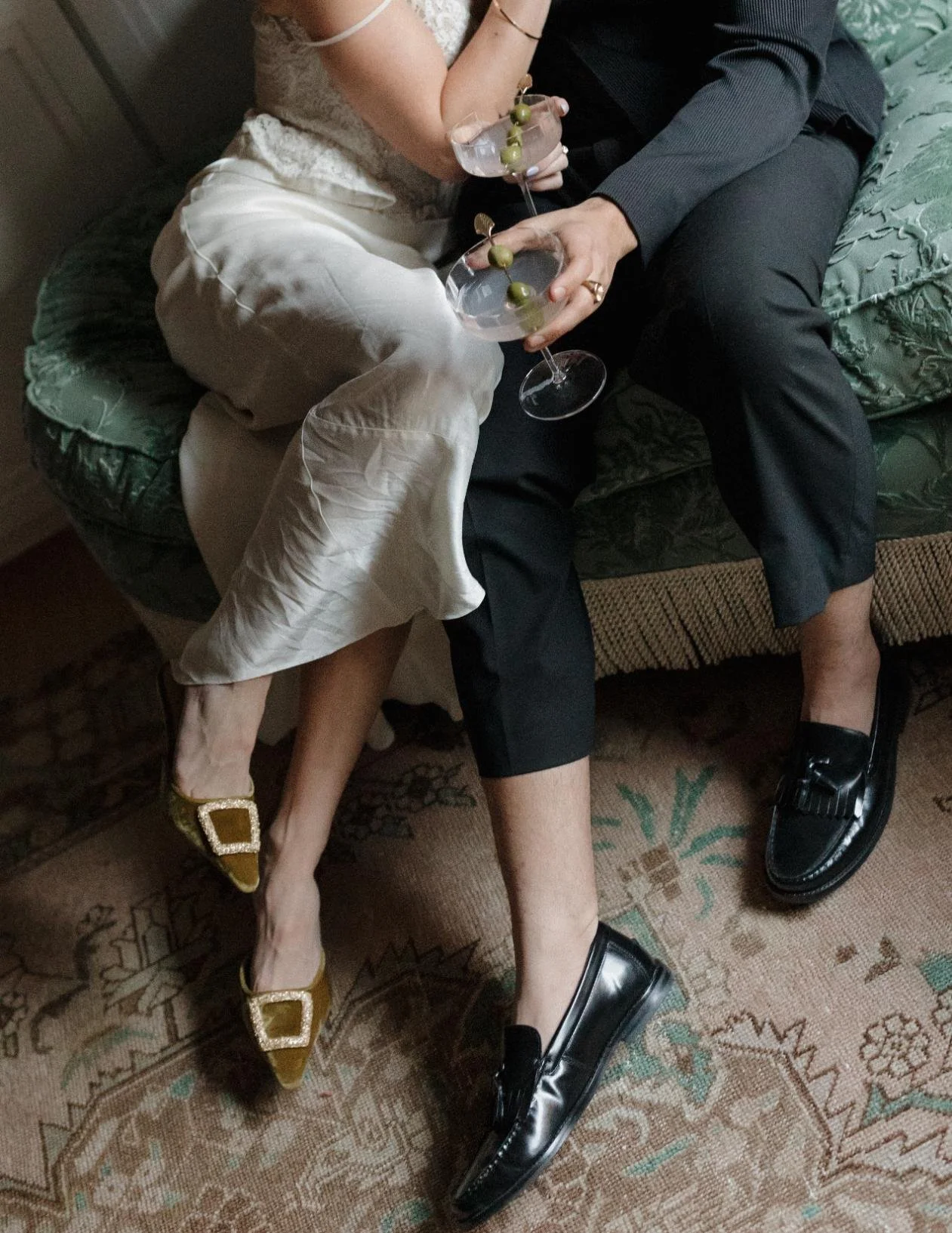 Two people sitting close together on a vintage-style green sofa, holding cocktail glasses with green olives, dressed in stylish evening attire. One person wears shiny black loafers; the other wears gold shoes with large decorative buckles.