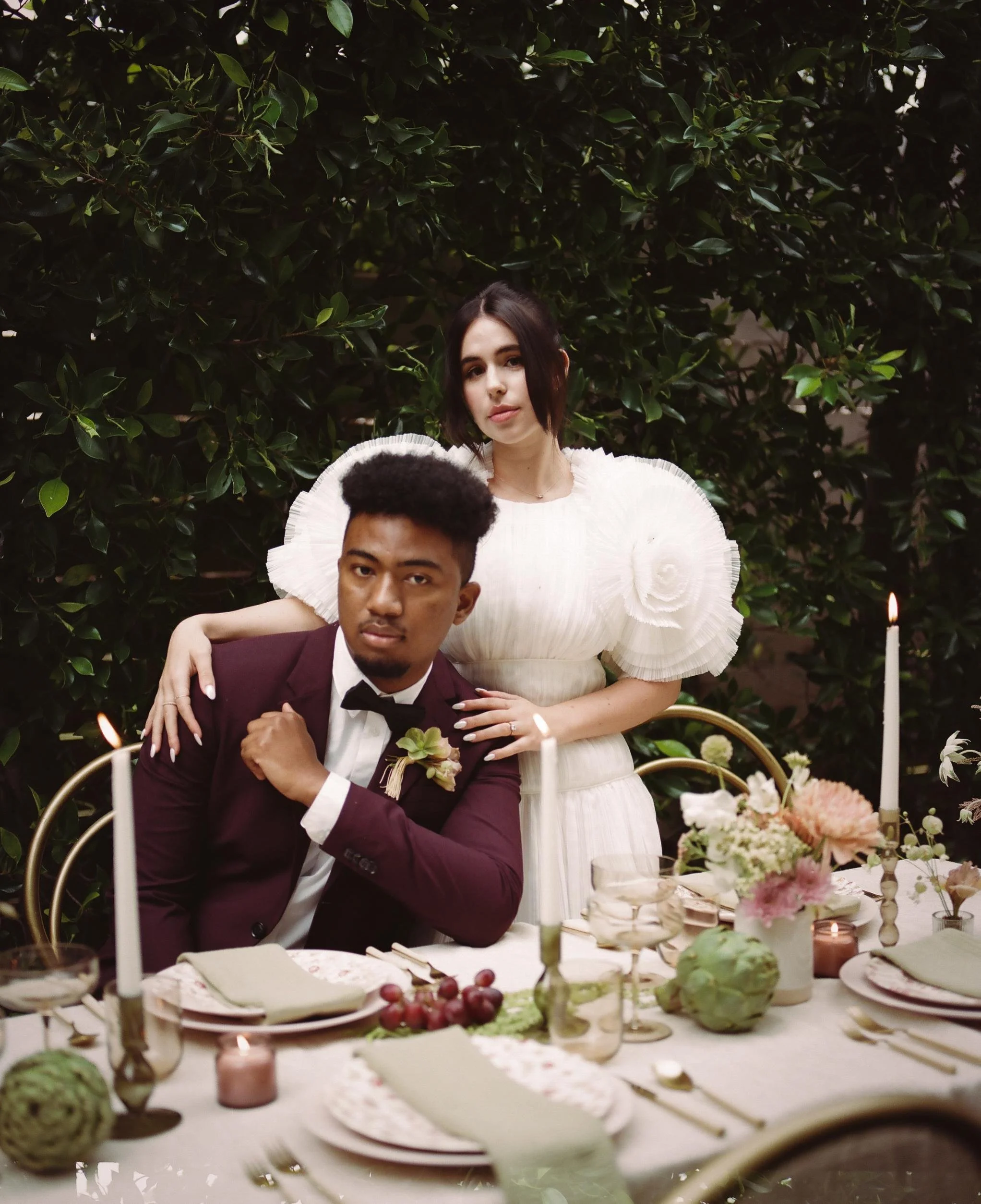 A young man in a burgundy tuxedo and a young woman in a white dress are posing at a wedding reception table surrounded by candles, flowers, and greenery.