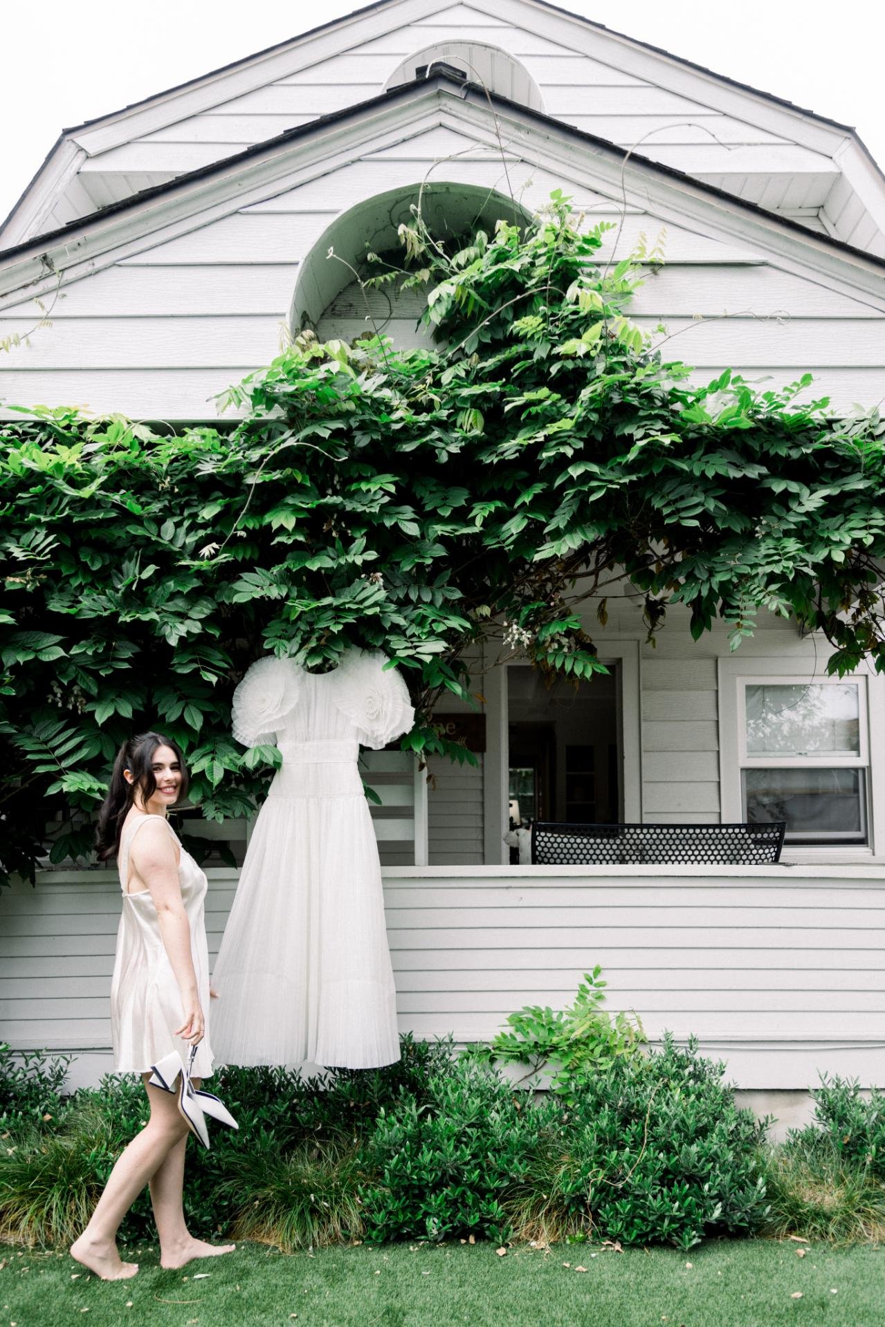 A woman smiling and standing barefoot on grass in front of a house with white siding, holding a pair of white shoes, with a large green leafy vine covering part of the house and a hanging white dress sculpture.