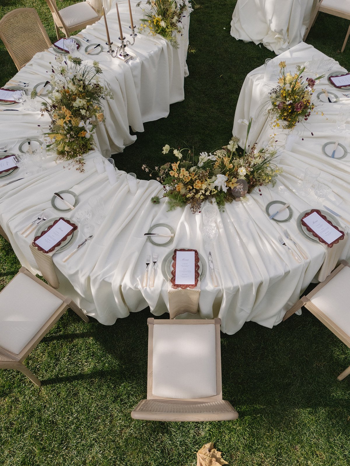 A decorated outdoor banquet table with a white tablecloth, floral centerpieces, place settings, and chairs arranged in a semi-circle on grass.