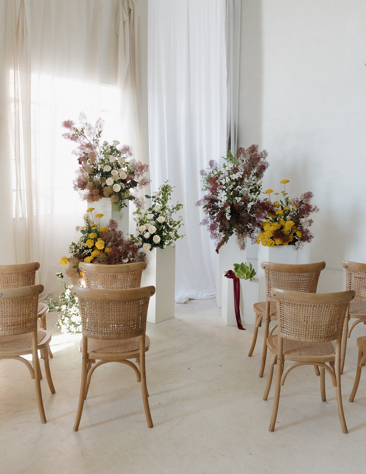Indoor setting with beige wicker chairs and flower arrangements on white pedestals near sheer curtains and bright natural light