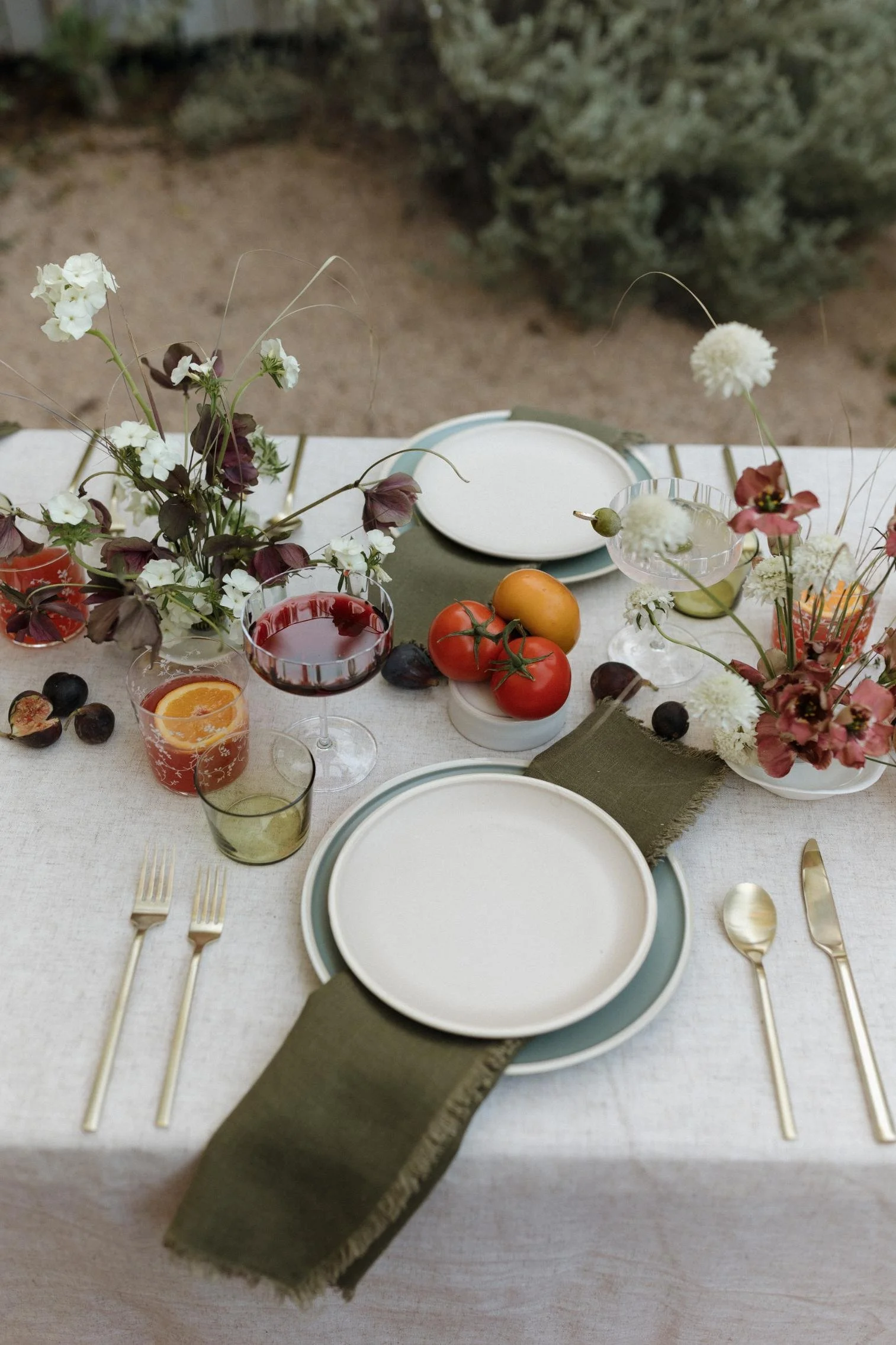 A beautifully set outdoor dining table with white plates, gold utensils, and floral centerpieces, decorated with fresh tomatoes, figs, wine glasses, and candles.