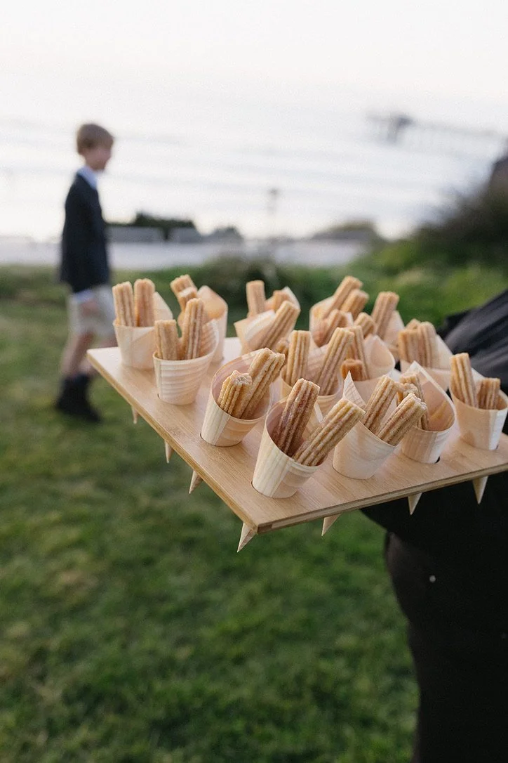 Person holding a wooden tray with churros in small paper cups outdoors, with a boy in a blazer walking in the background.