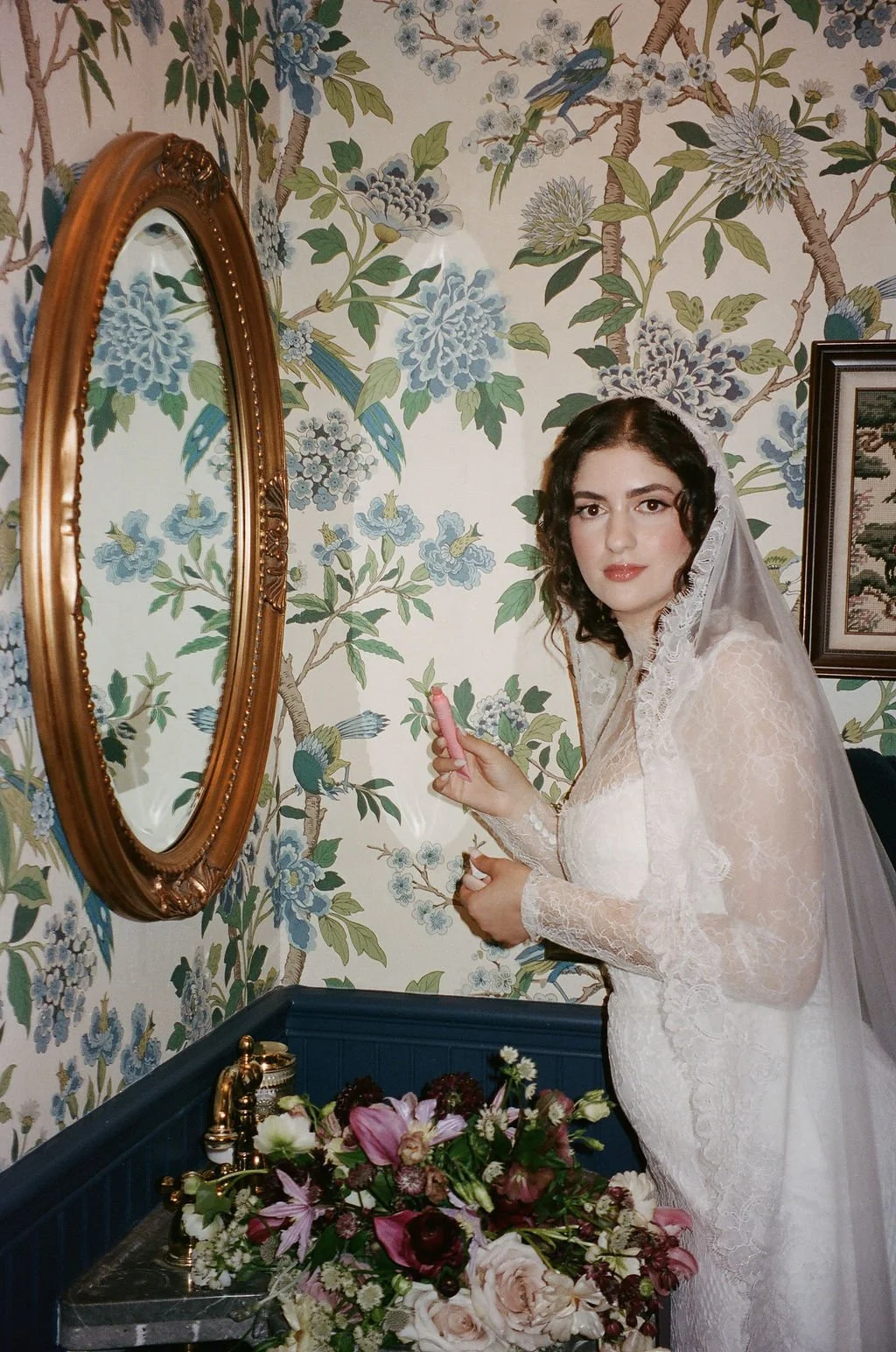 A bride with dark curly hair, wearing a lace wedding dress and veil, holding a pink lipstick, in front of floral wallpaper with birds, standing beside a floral arrangement on a blue table.