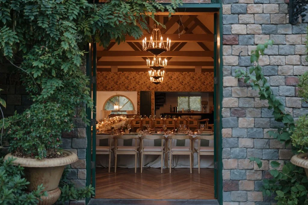 View of a decorated indoor event space seen through an open doorway framed by greenery, with chandeliers hanging from the ceiling, a long dining table, and windows in the background.