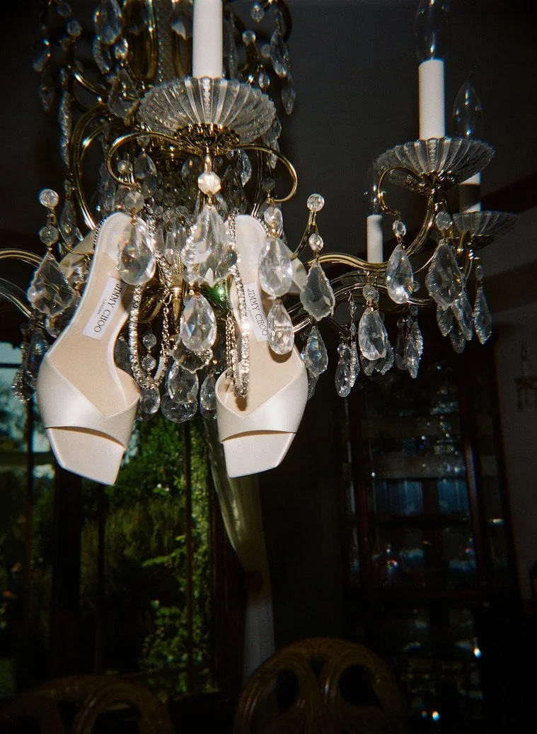 A chandelier hanging from the ceiling with crystal and glass decorations. A pair of white Jimmy Choo high-heeled shoes are also hanging from the chandelier.