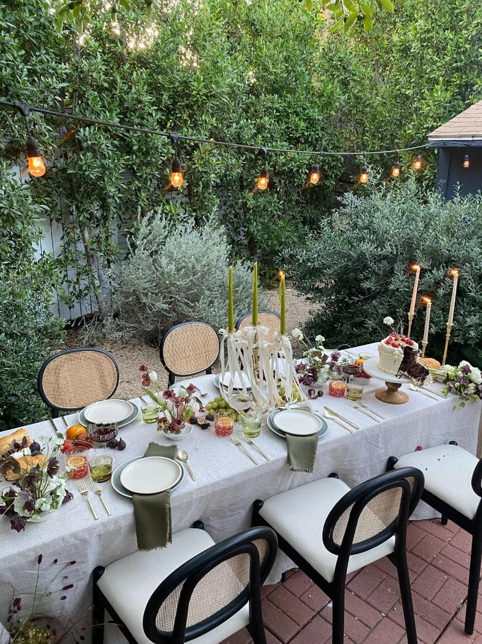Outdoor dining table decorated with a white tablecloth, floral arrangements, and candles, set for a celebration with chairs around it, string lights overhead, surrounded by greenery.