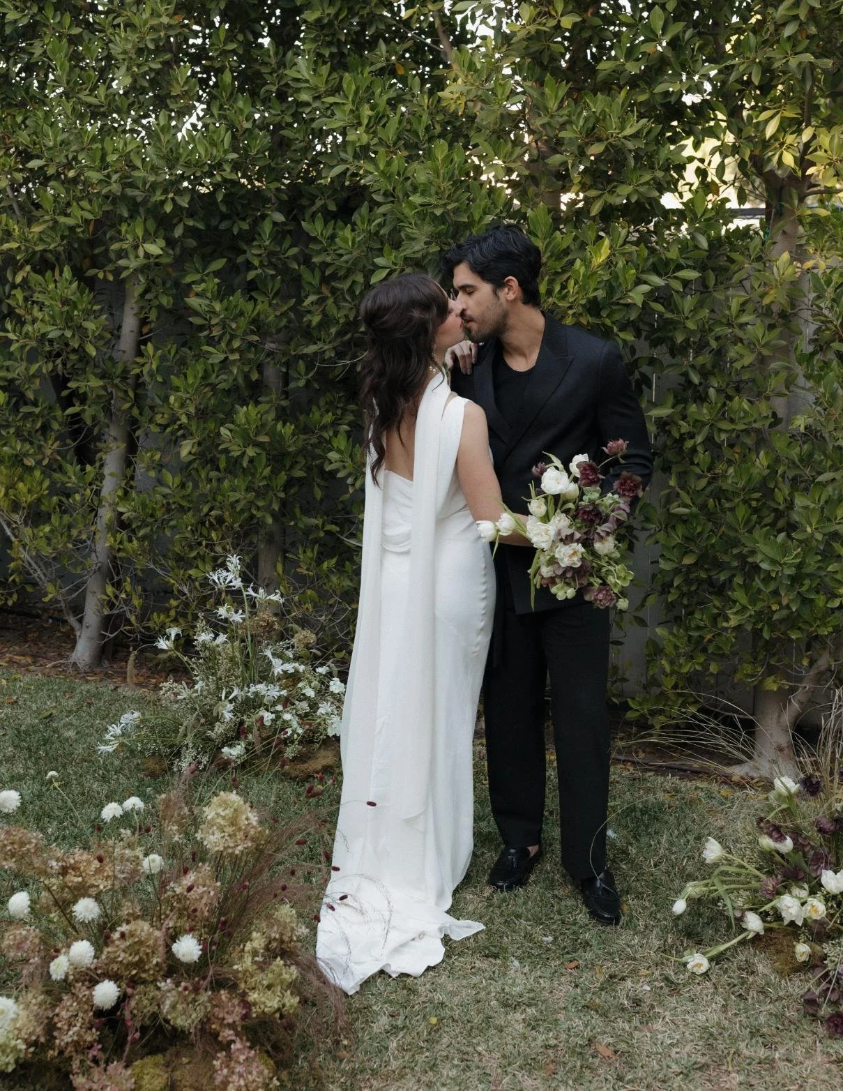 A bride and groom sharing a kiss outdoors, standing on grass with green bushes behind them. The bride wears a white gown with floral accents, and the groom wears a black suit. The groom holds a bouquet of flowers.