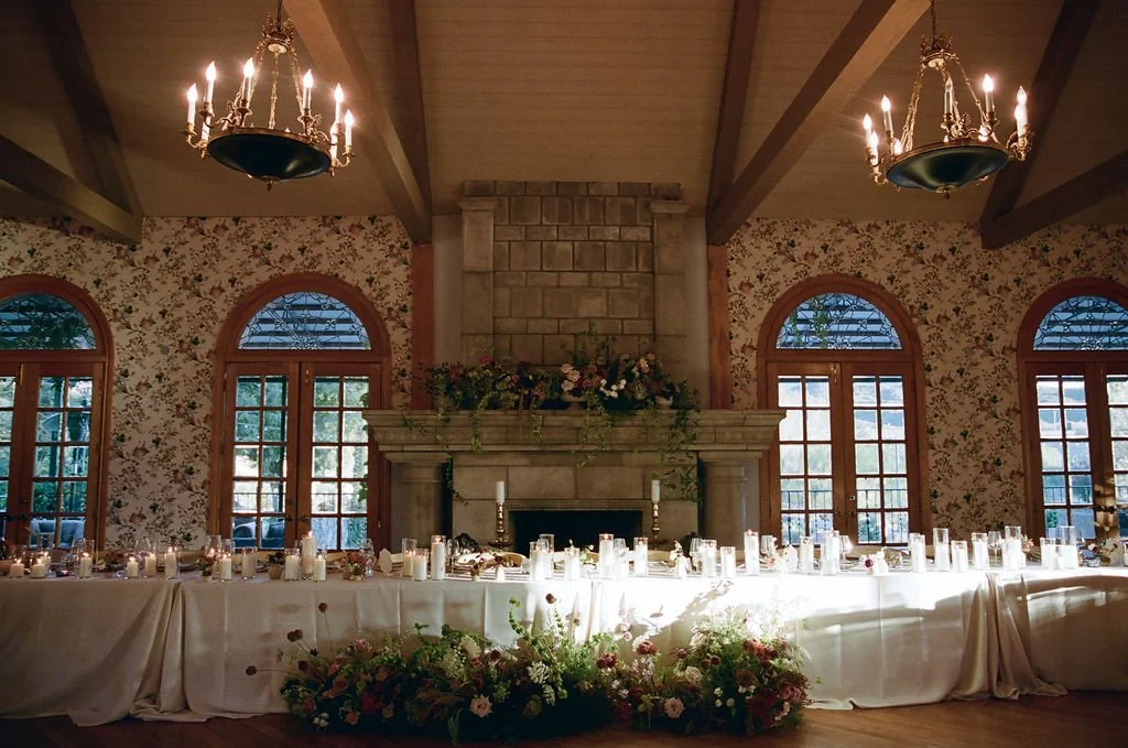 A long banquet table decorated with candles and floral arrangements in a rustic room with wooden beams, floral wallpaper, a stone fireplace, and large arched windows with sunlight.