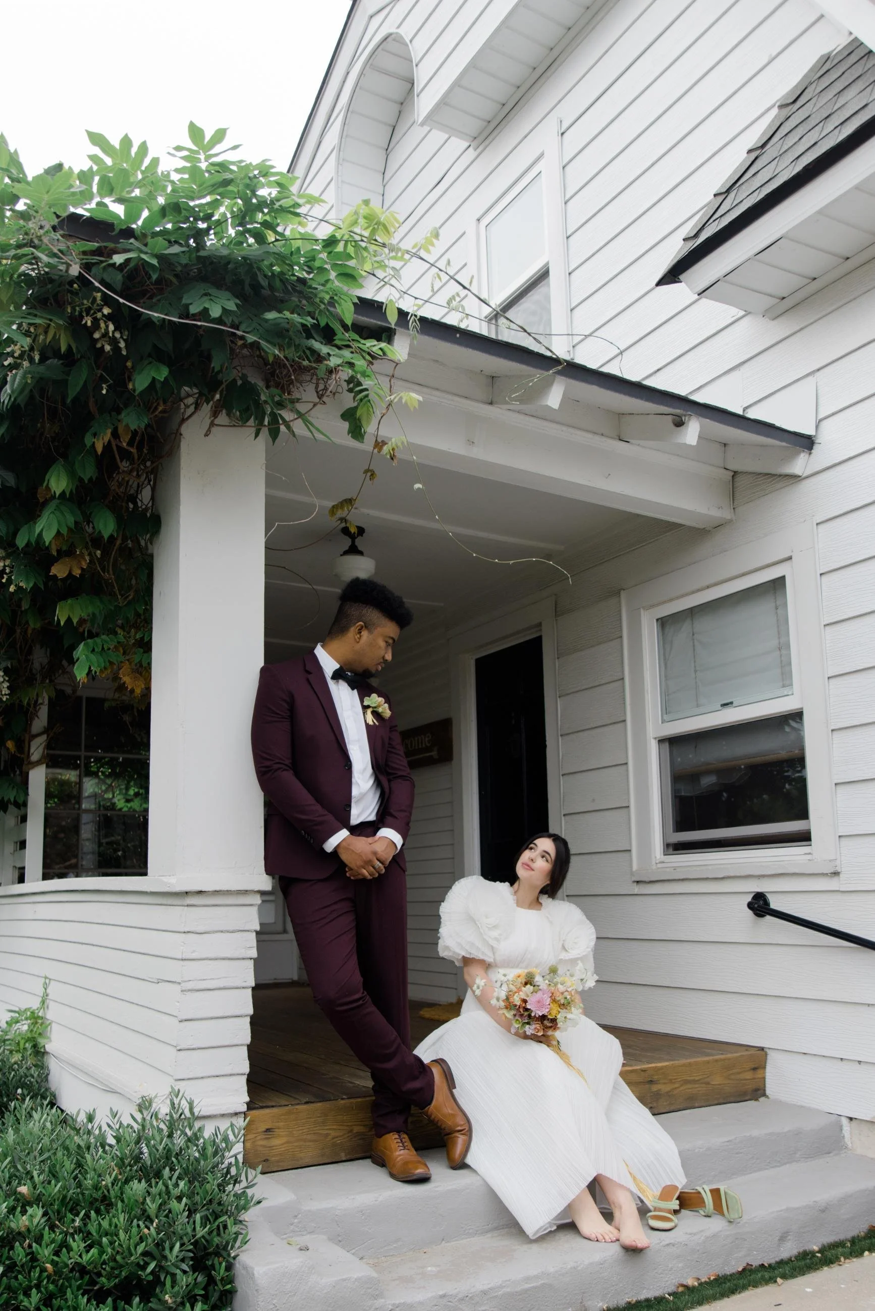A bride sitting on steps holding a bouquet, looking up at a groom standing next to her, on a porch with white siding and greenery.