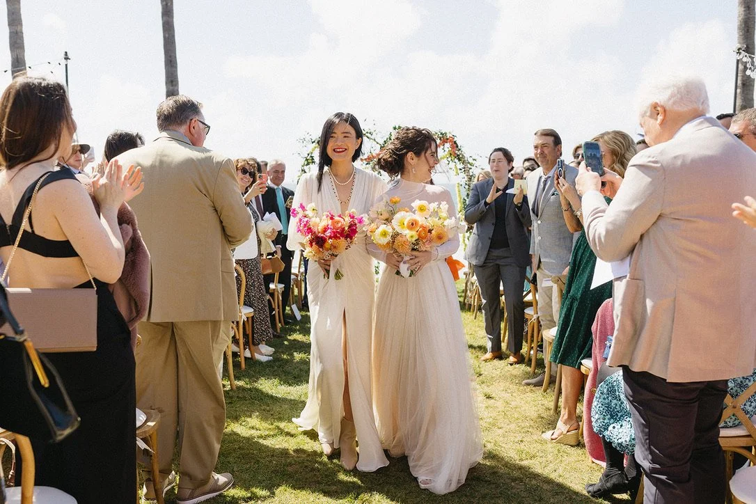 Two women in wedding dresses walking down the aisle at a wedding ceremony, holding bouquets of flowers, surrounded by seated guests taking pictures and applauding outside on a grassy area.