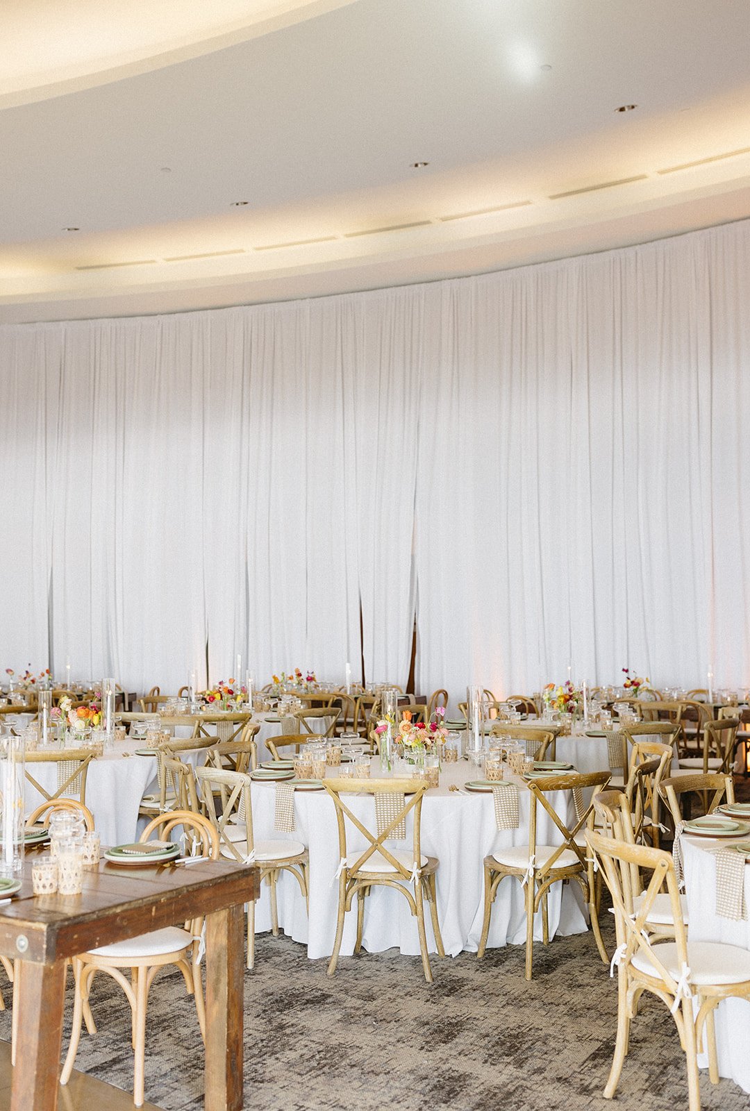 Event setup with multiple round tables covered in white tablecloths, decorated with floral centerpieces, candles, and tableware, against a backdrop of white curtains in a banquet hall.