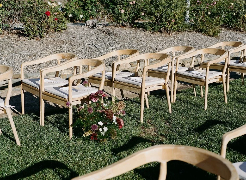 Several wooden chairs with white cushions arranged outdoors on grass for an event, with a flower arrangement and rose bushes in the background.