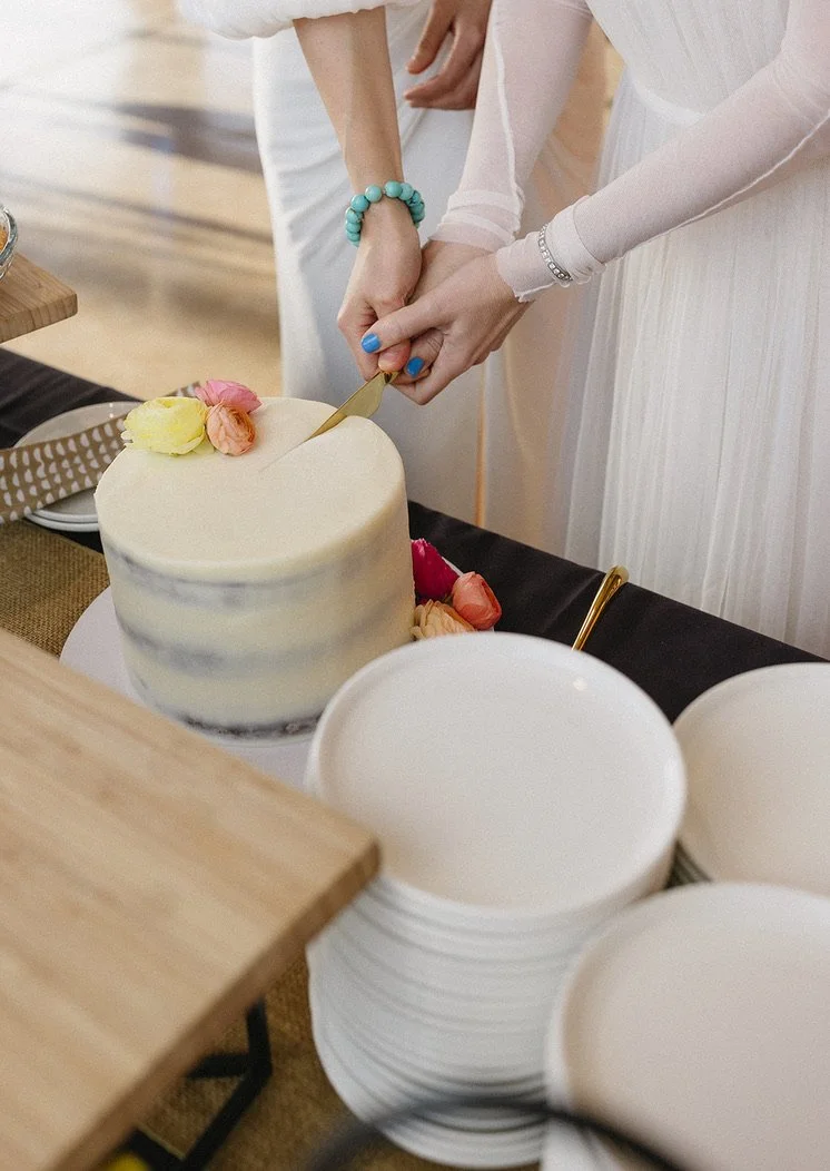 Two people cutting a wedding cake together at a celebration, with plates and utensils nearby.