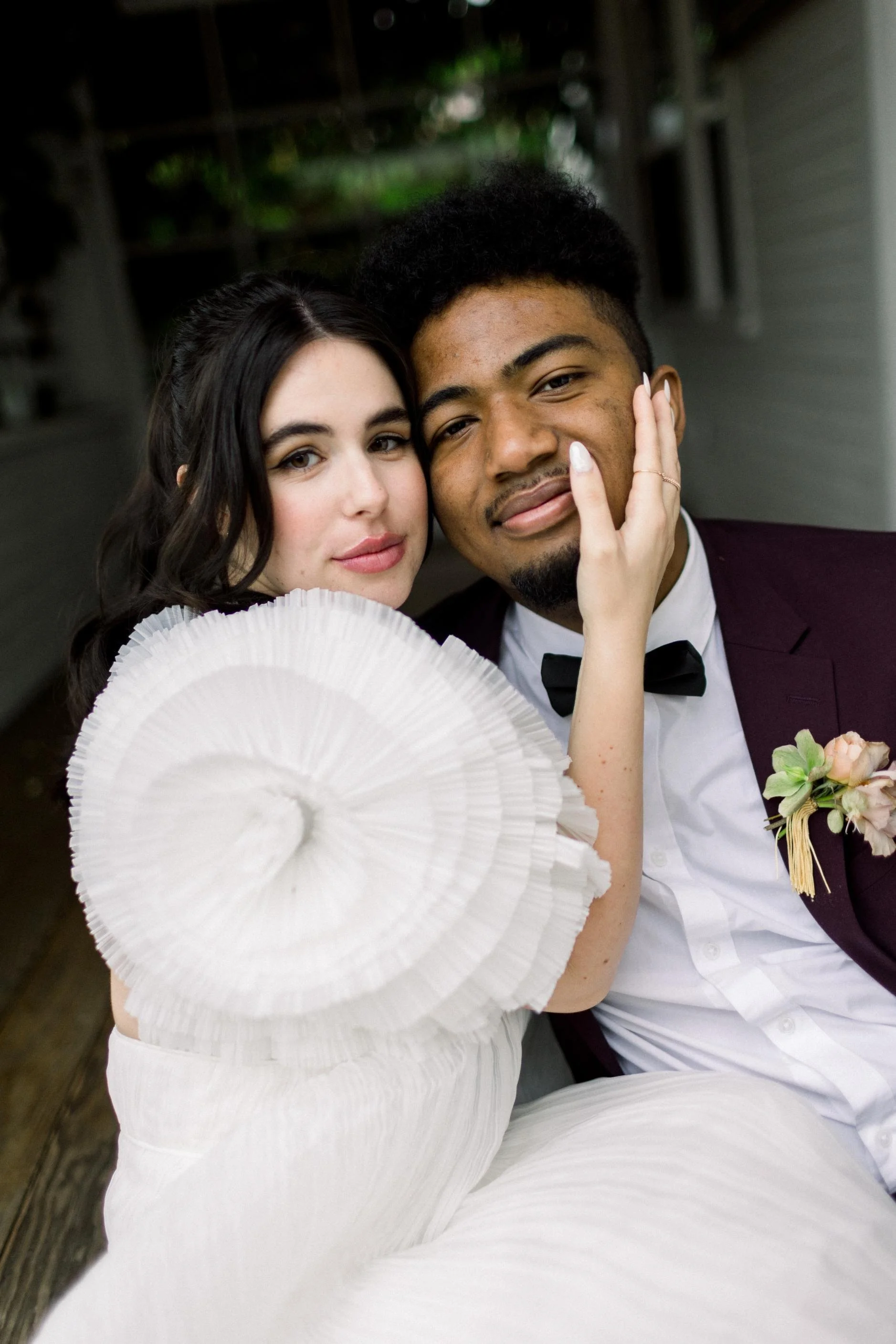 A bride and groom at their wedding, embracing closely, with the bride touching the groom's face. The bride is holding a white paper parasol and wears a white dress with a ruffled sleeve. The groom is dressed in a burgundy tuxedo with a white shirt an