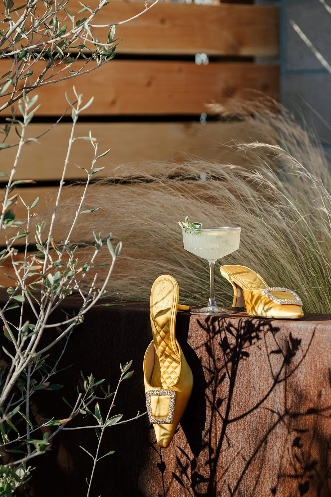 A cocktail glass with a drink and a small garnish, a pair of gold high-heeled shoes with rhinestone embellishments, placed on a stone surface outdoors with plants and shadows.