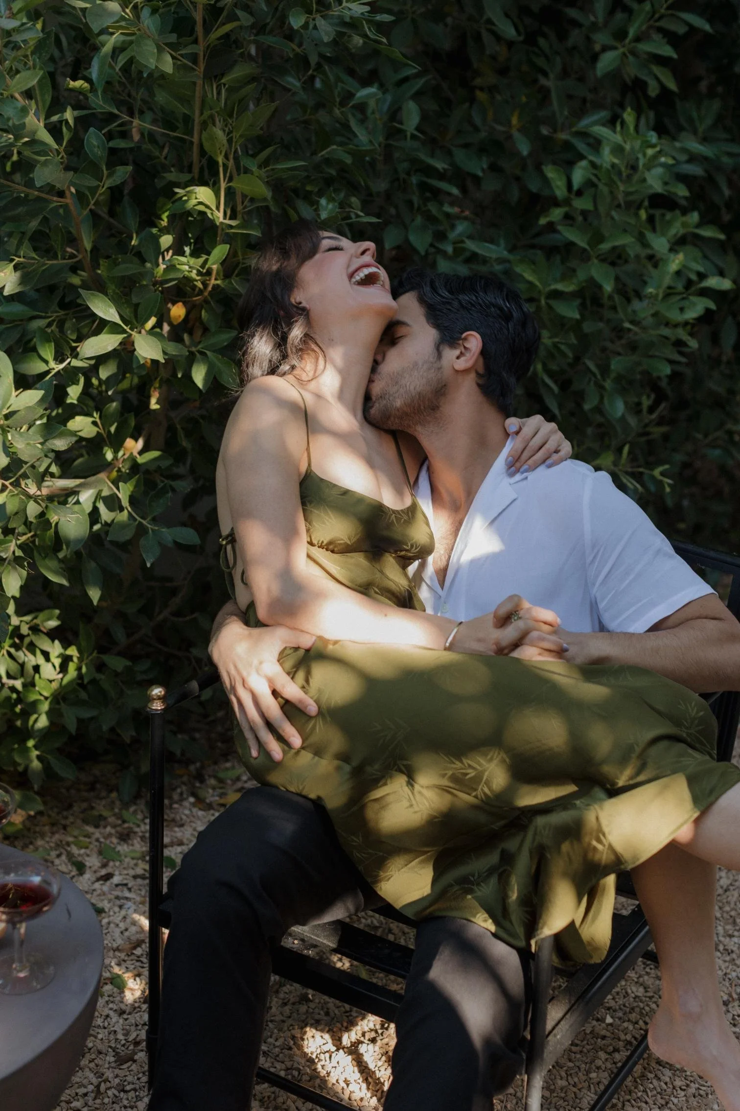 A couple sitting together outdoors, with the woman laughing and the man kissing her cheek, surrounded by lush green foliage.