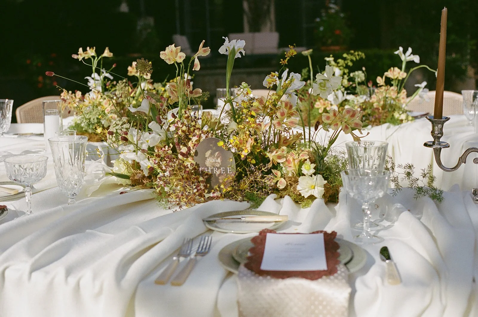Elegant wedding or event table decorated with a large floral centerpiece composed of white, light pink, and cream-colored flowers, with glassware, plates, and silverware arranged around it, and a candelabra with a single unlit taper candle on the right side.