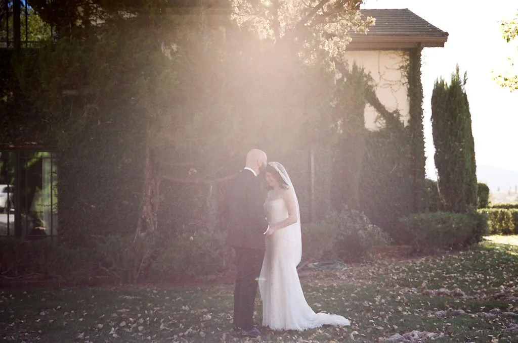 A bride and groom standing close together outdoors during a wedding ceremony, with sunlight shining overhead, surrounded by trees and a house in the background.