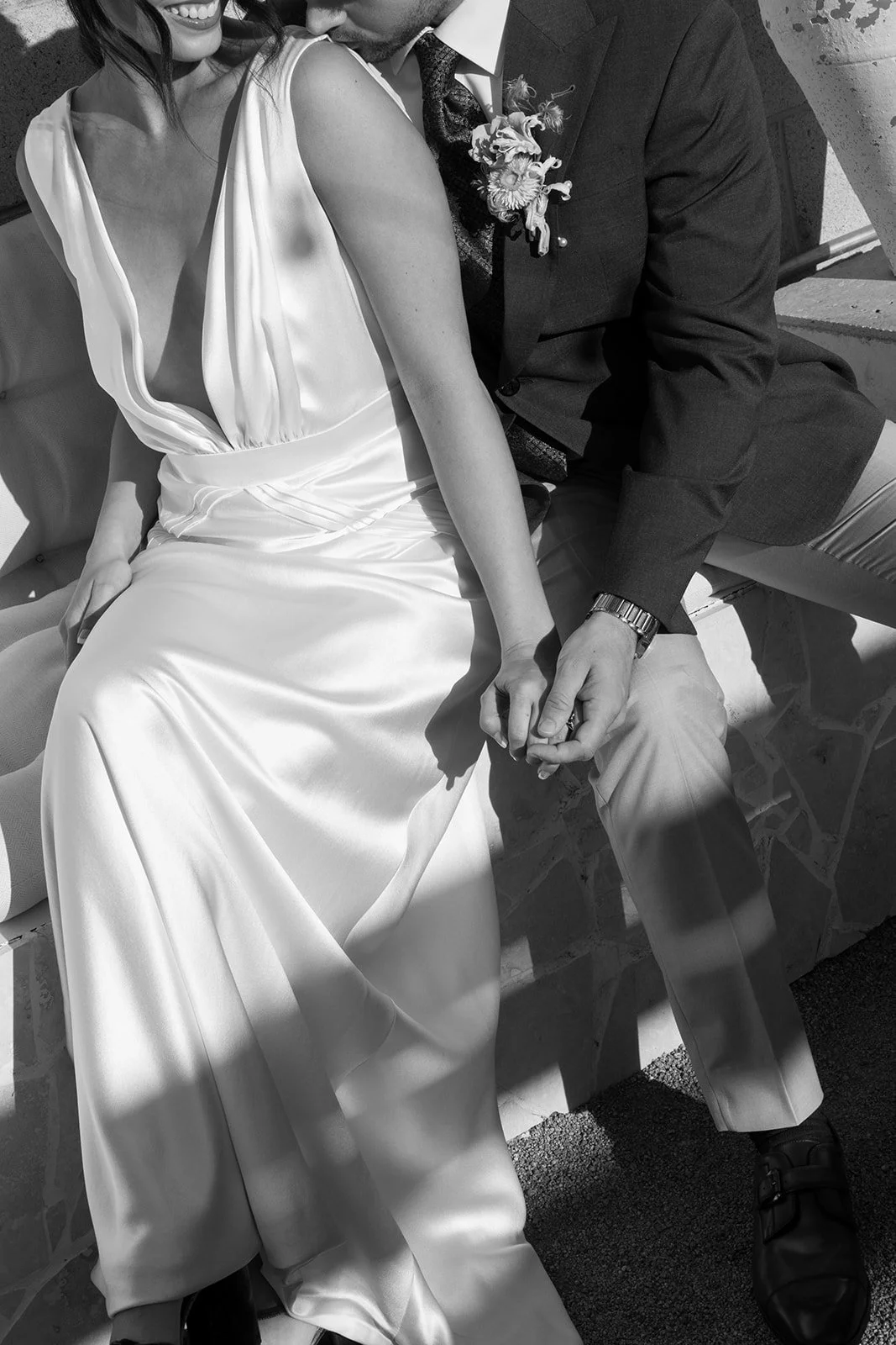 A close-up black and white photo of a couple holding hands, with the woman in a satin dress and the man in a suit, sitting on a stone bench.