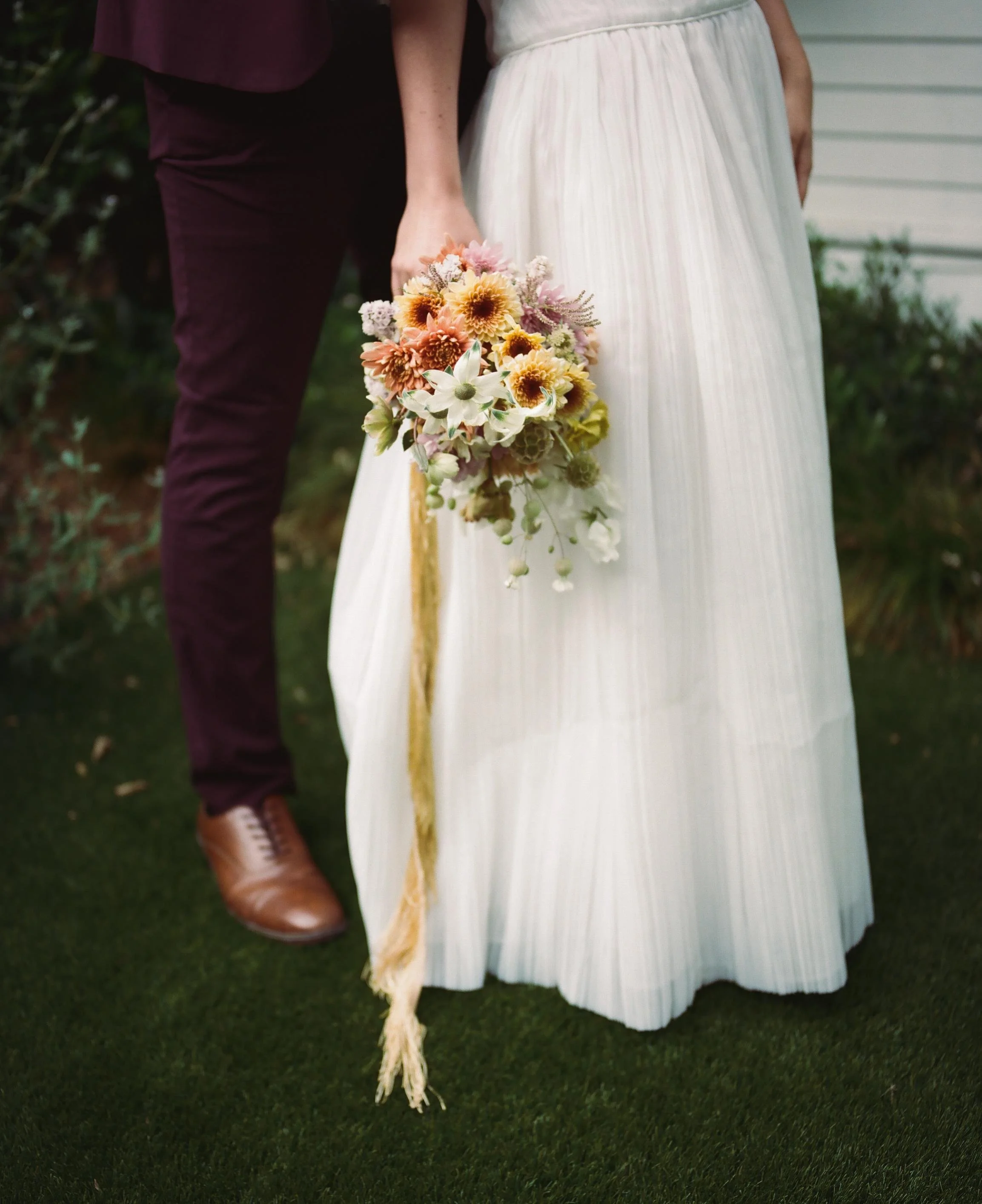 Partially visible couple holding a colorful bouquet of flowers outdoors, woman in a white pleated dress and man in dark pants and brown shoes.
