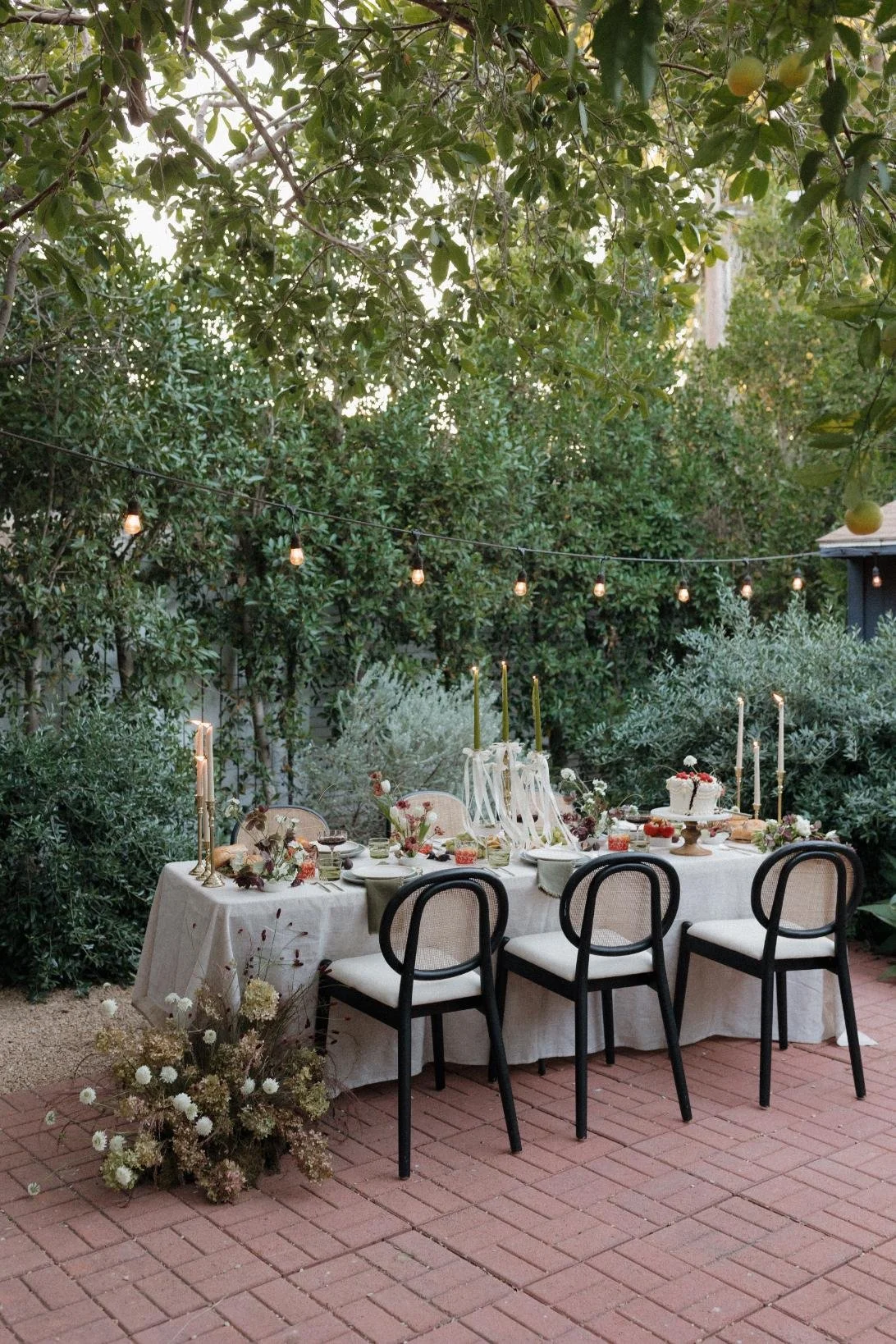 An outdoor dining setup on a patio with a white tablecloth, decorated with candles, flowers, and tableware, surrounded by greenery and string lights overhead.
