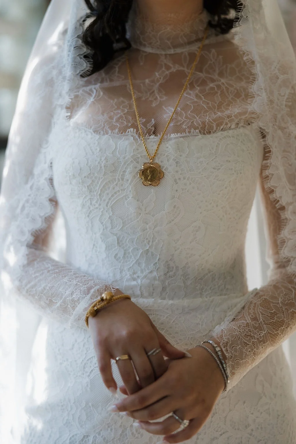 Close-up of a woman dressed in a white lace dress with a high neckline, wearing layered jewelry including a gold chain with a flower pendant, bracelets, and rings, with dark curly hair.
