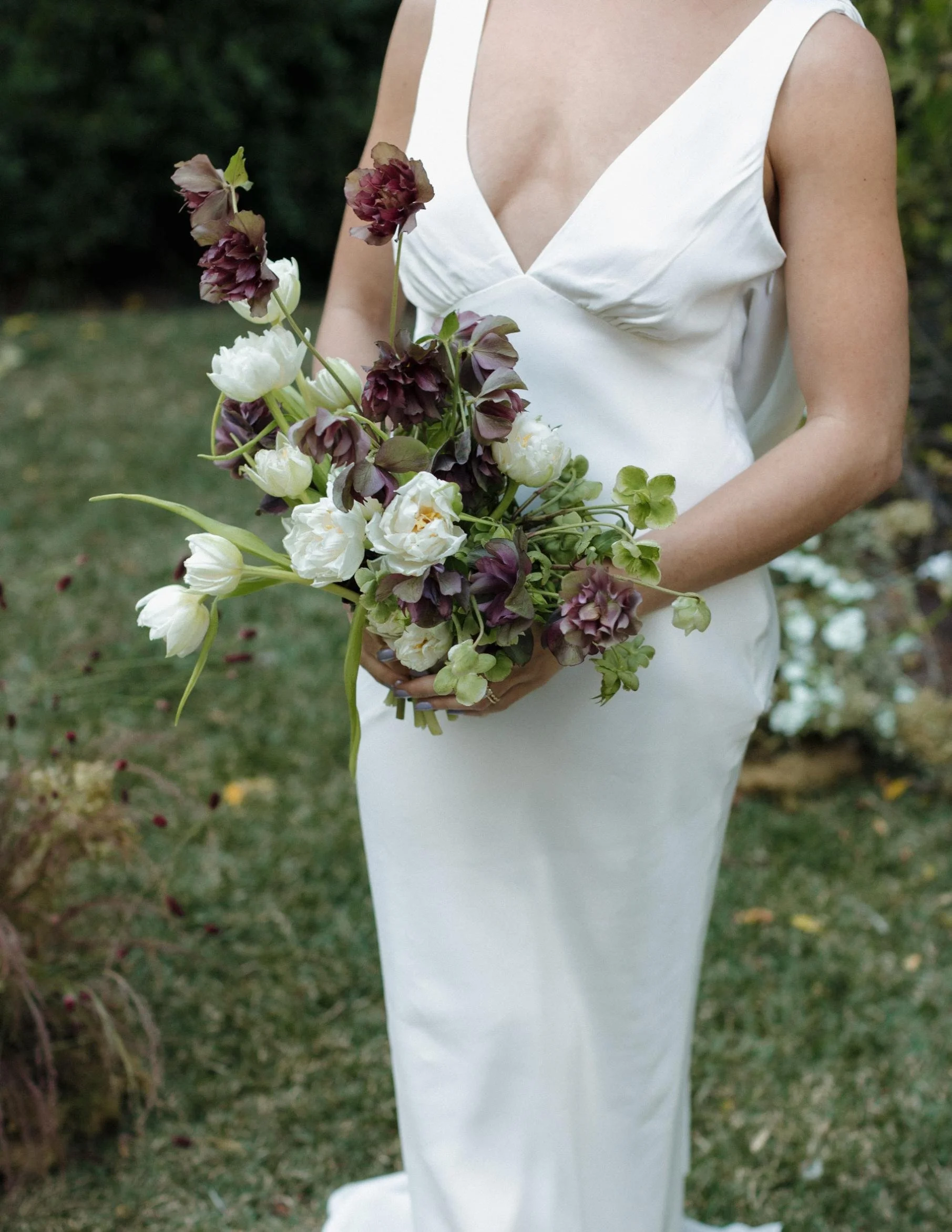 A woman in a white dress holding a bouquet of dark purple and white flowers outdoors.