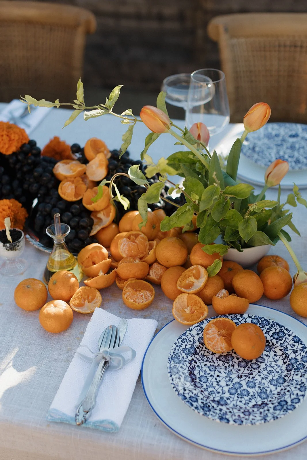 A table decorated with oranges, mandarin oranges, and a floral centerpiece. There are blue and white patterned plates, water glasses, and silverware wrapped in a white napkin with a ribbon.