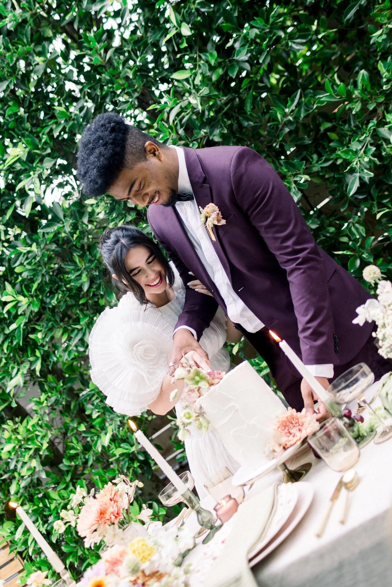 A bride and groom are cutting a wedding cake together during their celebration, standing at a decorated table with flowers and candles.