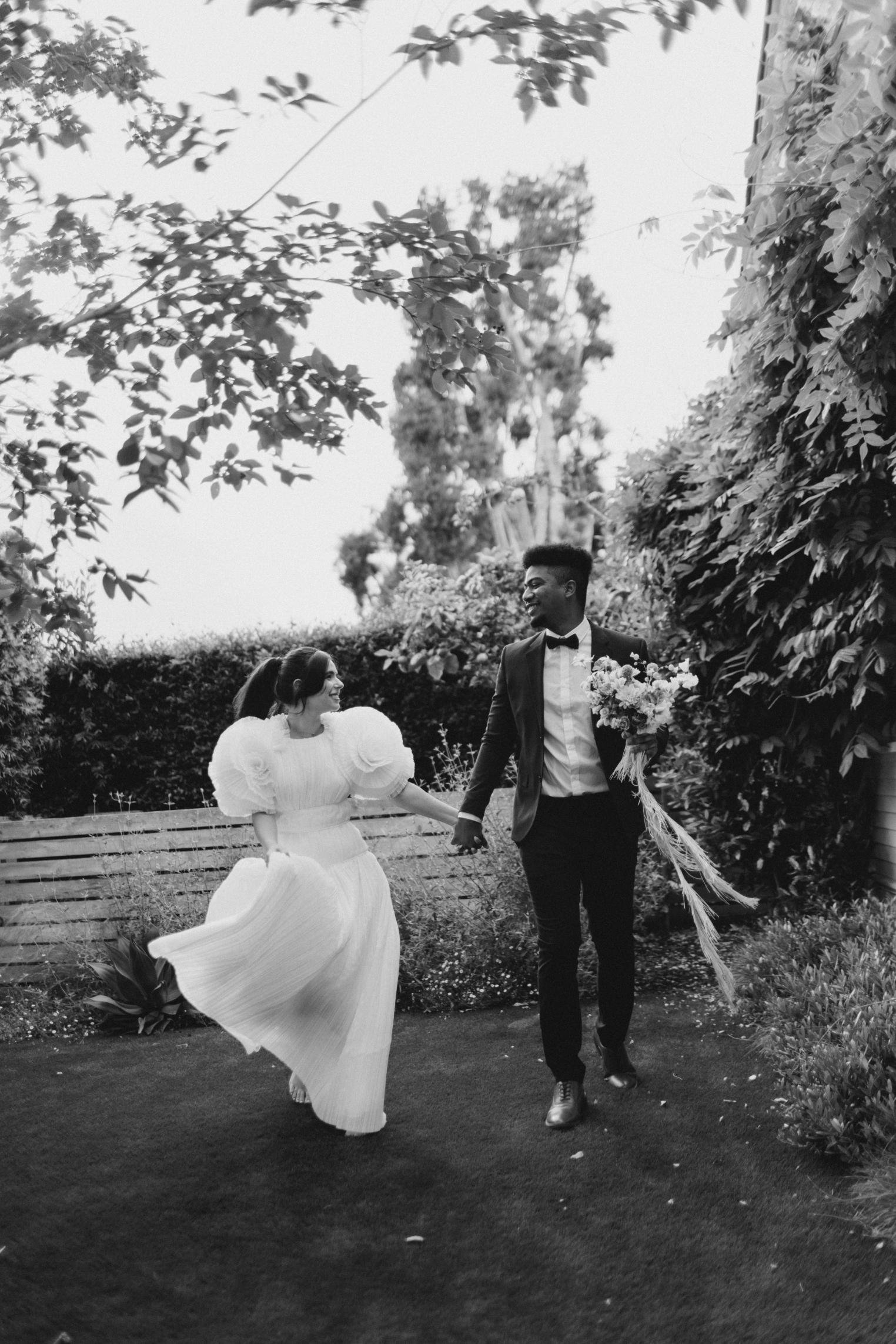 A black-and-white photo of a wedding couple holding hands and walking in a garden. The woman wears a white gown with puffed sleeves and the man is in a suit with a bowtie, holding a bouquet of flowers.
