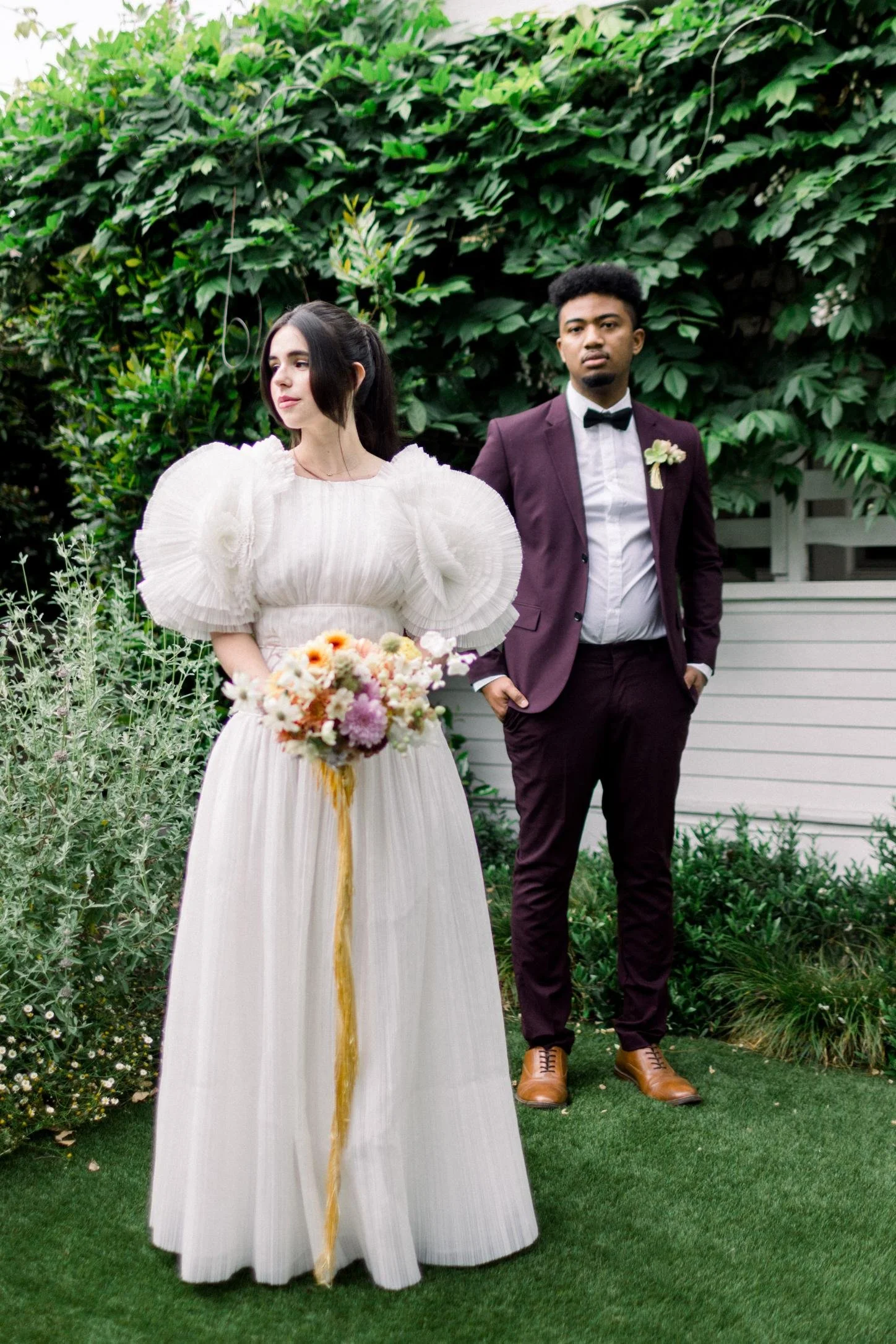 A woman in an elegant white dress with large puffed sleeves holds a bouquet of flowers. A man in a dark burgundy suit, white shirt, and black bow tie stands behind her. They are outdoors with lush green foliage in the background.