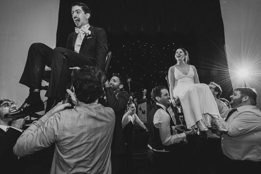Men are lifting women on chairs in a celebration, with one man on a chair and some holding another woman, in a lively event with a decorated stage in the background. Black and white photo.