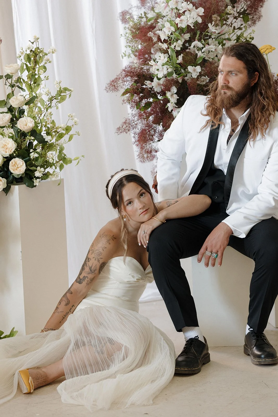A woman in a white wedding dress with tattoos leaning on the lap of a man in a white blazer and black pants, surrounded by flowers.