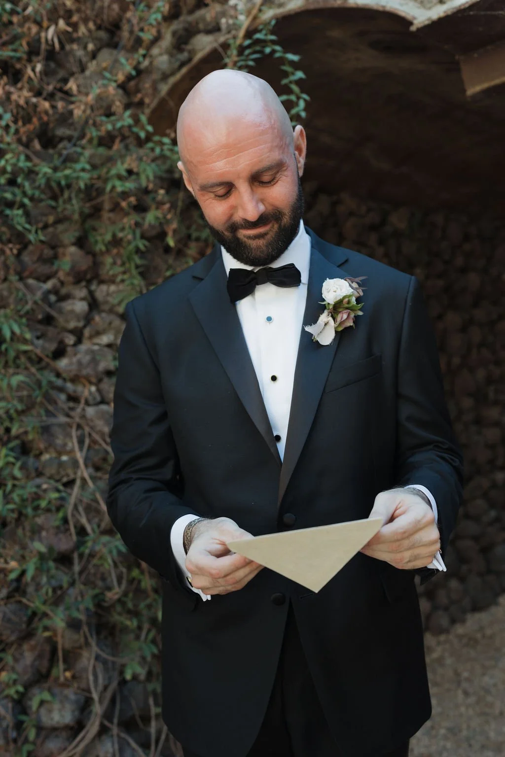 A man dressed in a black tuxedo with a white shirt and black bow tie, holding a checkered card, outdoors near a rocky area and greenery, with a smile and a boutonniere on his left lapel.