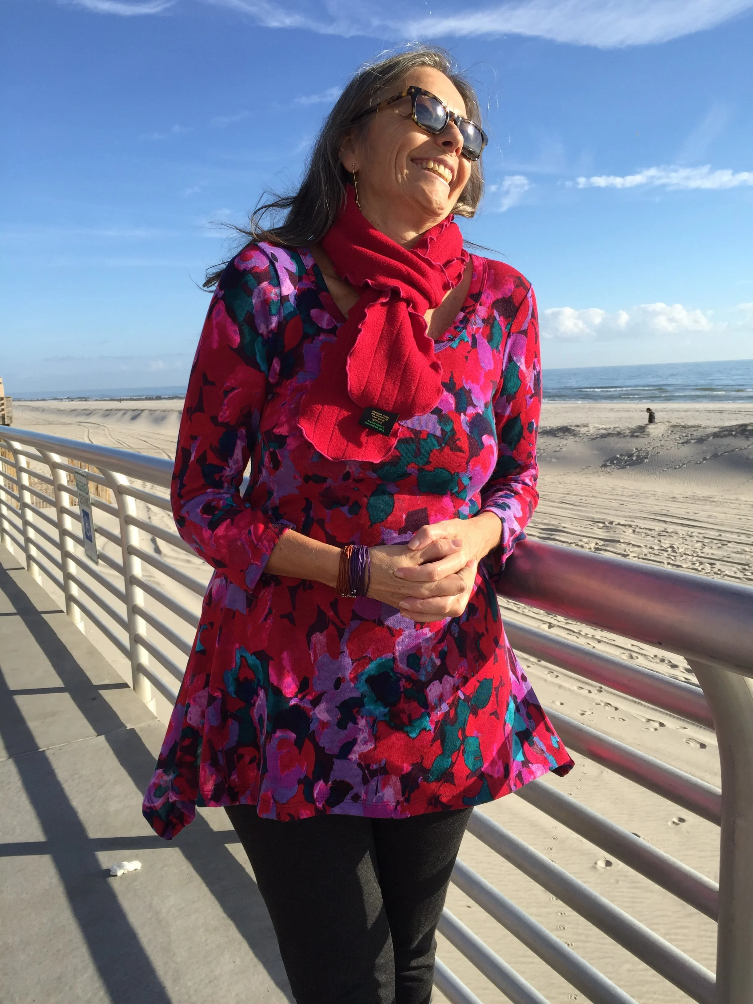 Woman in colorful floral top, red scarf, sunglasses, and black pants standing on a beach pier with photo of the ocean and sky in the background.