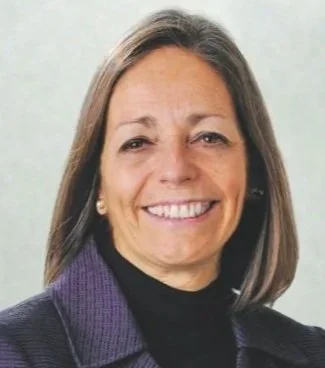 Close-up of a middle-aged woman with shoulder-length brown hair, smiling, wearing a purple blazer and black turtleneck.