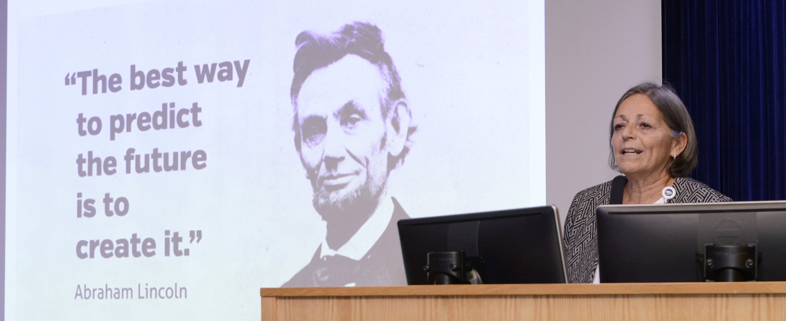 A woman standing at a podium giving a presentation. Behind her is a slide with a quote attributed to Abraham Lincoln that says, "The best way to predict the future is to create it," alongside a black-and-white portrait of Abraham Lincoln.
