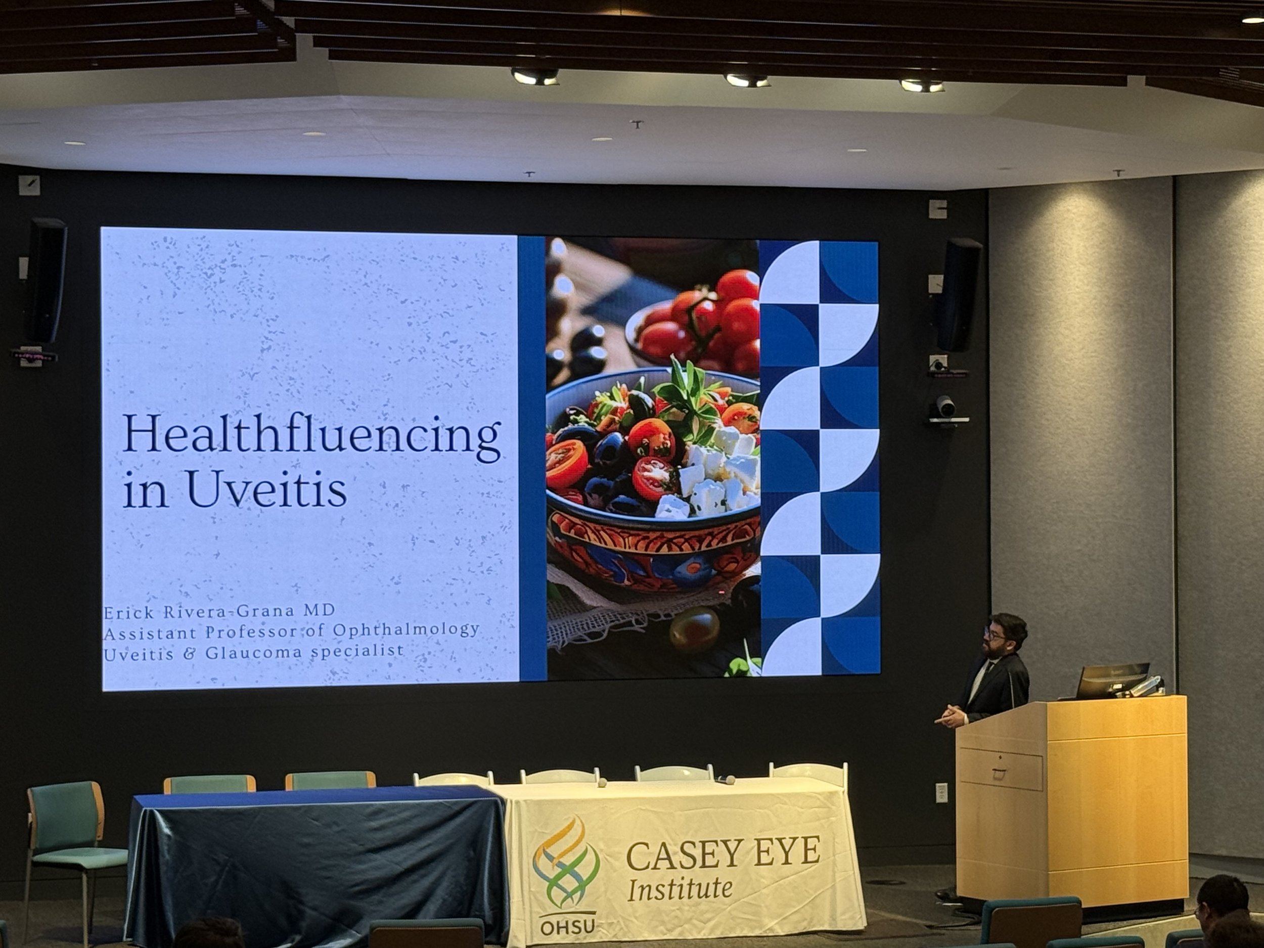A man giving a presentation on a large screen about health effects in uveitis. The slide shows a decorative blue and white pattern, a bowl of colorful salad, and the title 'Health influencing in Uveitis.' The presenter stands at a podium in a conference room with chairs and a table covered with a blue cloth and a banner that reads 'Casey Eye Institute'.