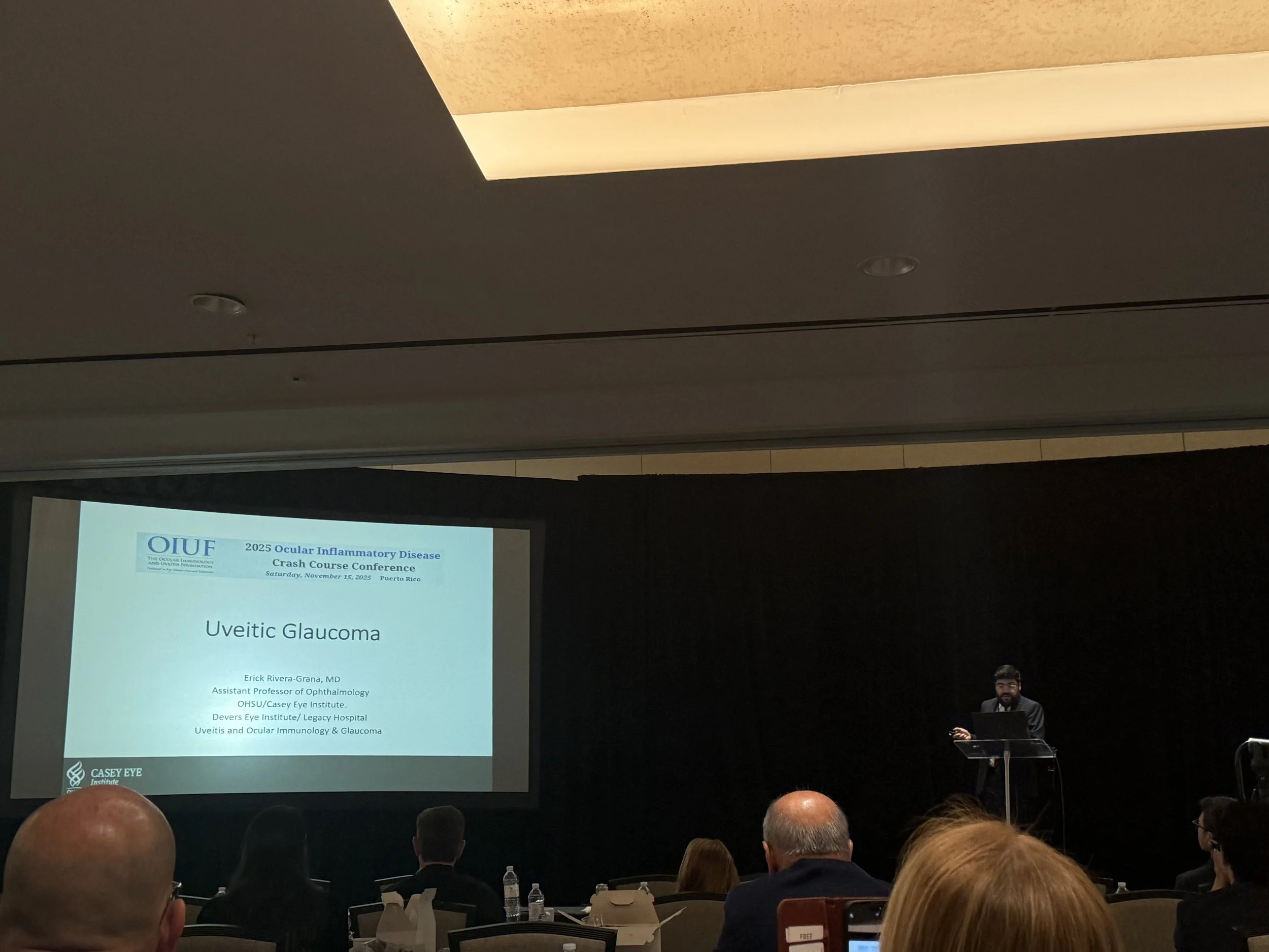 A medical conference presentation slide titled 'Uveitic Glaucoma' is projected on a screen. The slide includes the date November 25, 2023, and location Puerto Rico. A speaker stands at a podium on the right, with audience members seated and facing the screen.