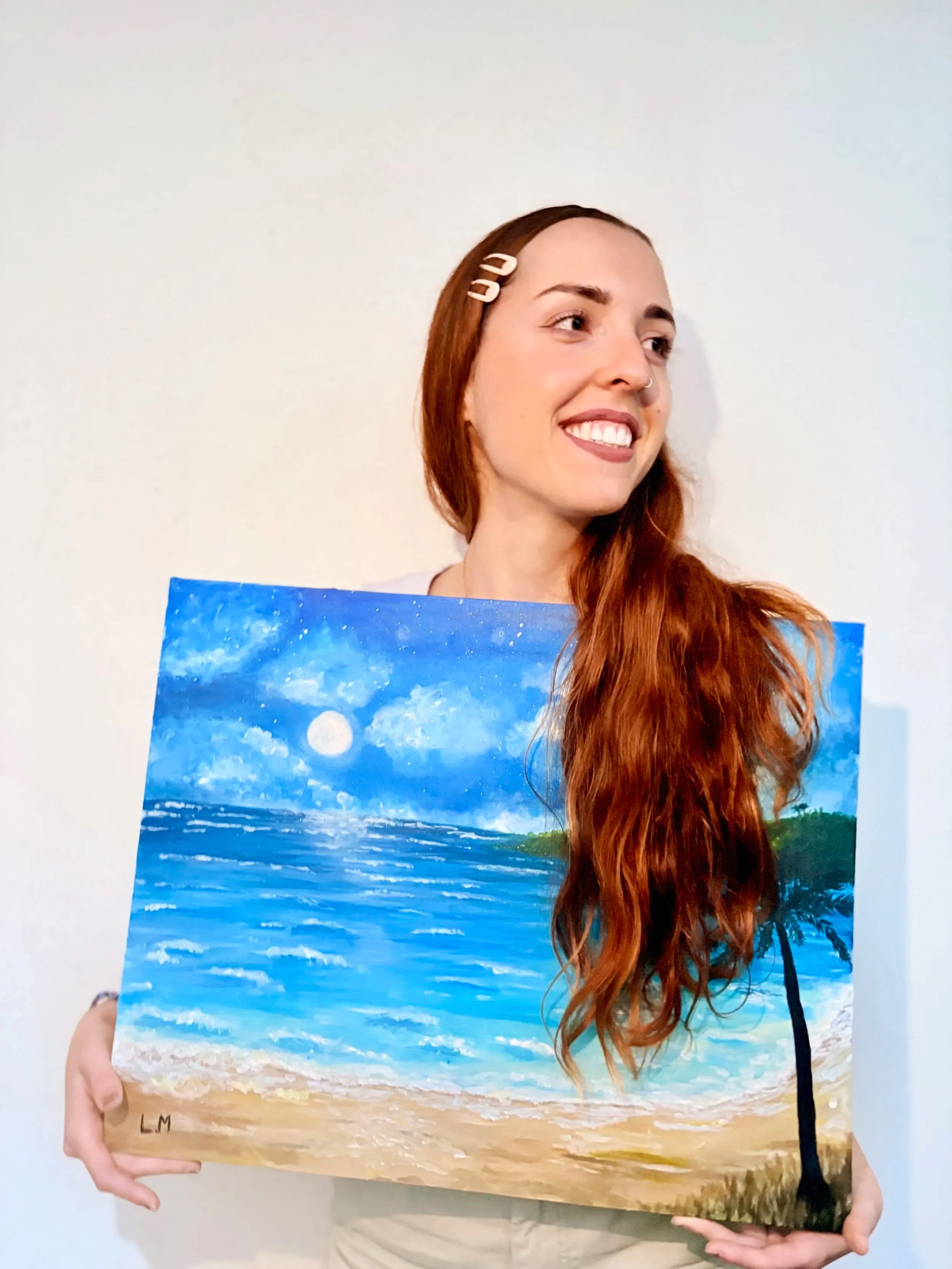 Une jeune femme rousse artiste LM PAINT tient un tableau peint à l'acrylique sur toile représentant une scène de plage avec la mer, le sable, une pleine lune et un ciel étoilé (Murmure du crépuscule par LM PAINT artiste peintre).