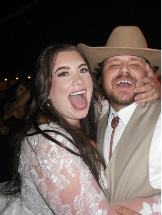 A woman and a man at a celebration, smiling and enjoying themselves. The woman has long dark hair and is wearing a white lace dress, and the man is wearing a beige cowboy hat and a western-style shirt with a vest.