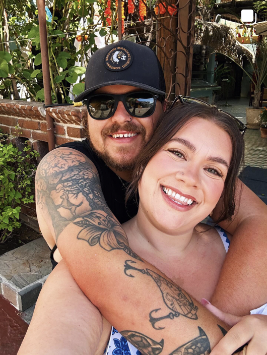 Happy couple taking a selfie outdoors, with the woman in front and the man hugging her from behind, both smiling broadly, in a garden setting.