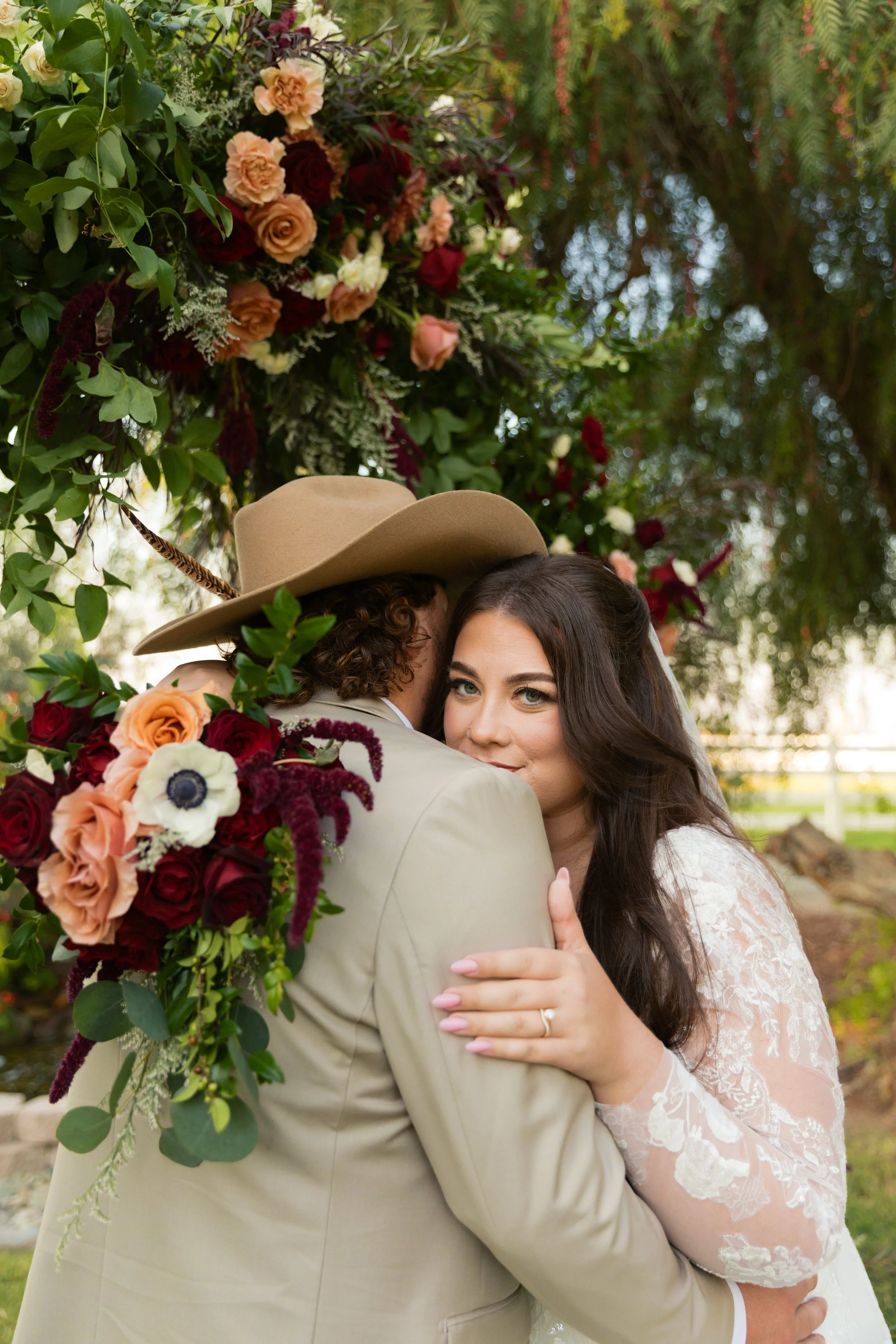 A couple hugging outdoors during a wedding, with the bride's face visible and the groom's face hidden. The bride has long dark hair and is wearing a lace wedding dress, while the groom is wearing a light beige suit and a wide-brimmed hat. They are standing under a floral arch decorated with red, peach, and white roses and greenery.