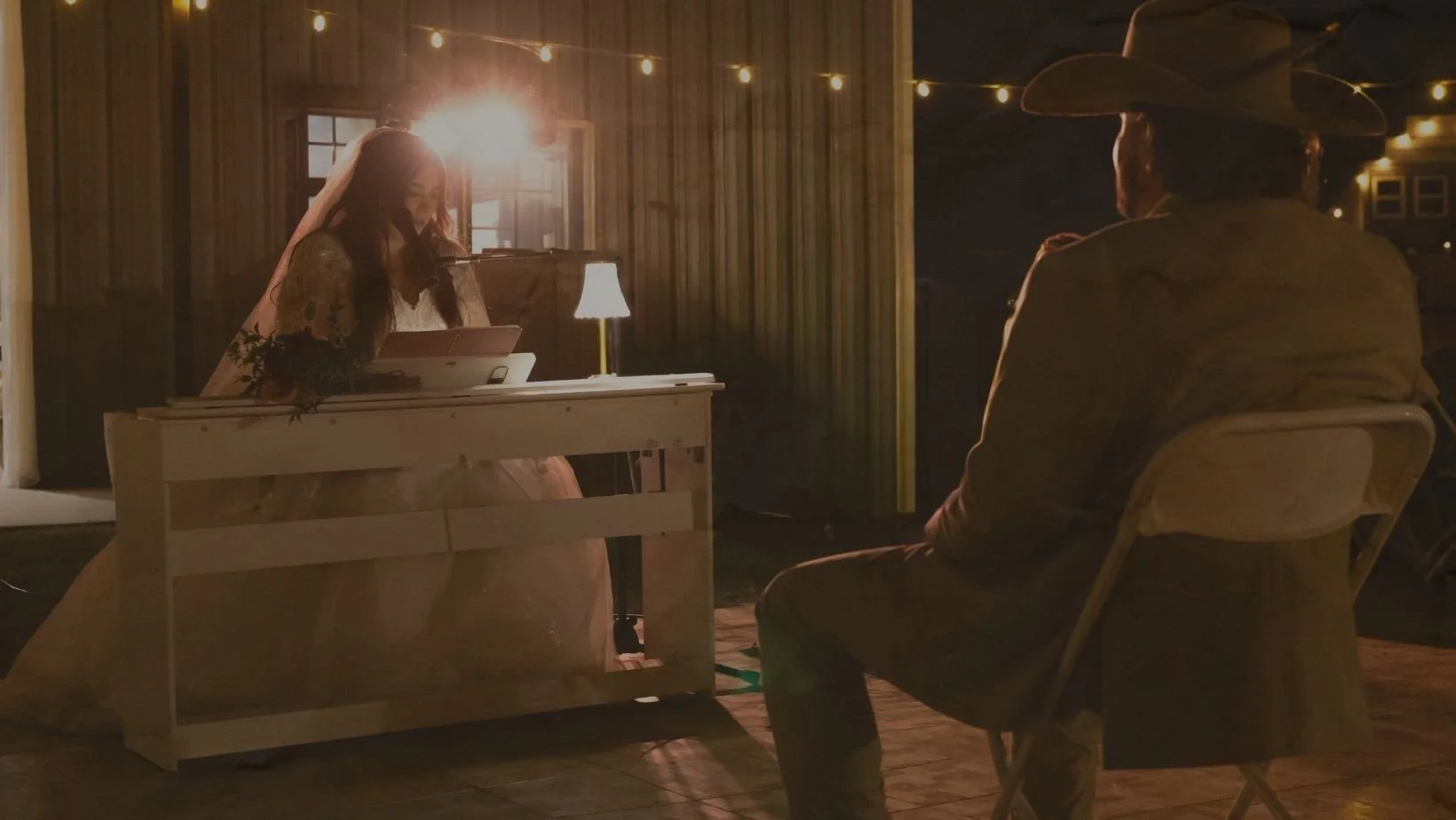 A woman in a wedding dress and a veil is playing a keyboard, smiling, while a man in a cowboy hat and western attire sits on a chair facing her outdoors during the evening, with string lights and a wooden structure in the background.