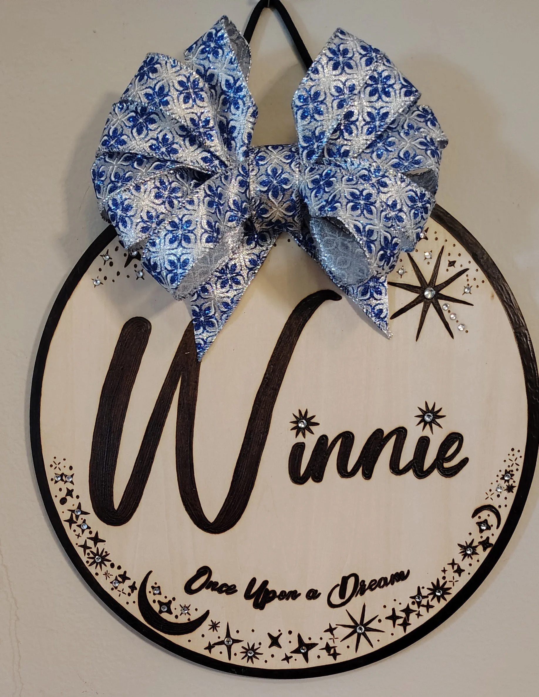 Decorative wall sign with a large blue and silver bow on top. The sign says "Winnie" with the subtitle "Once Upon a Dream" and is decorated with stars, moons, and sparkles.