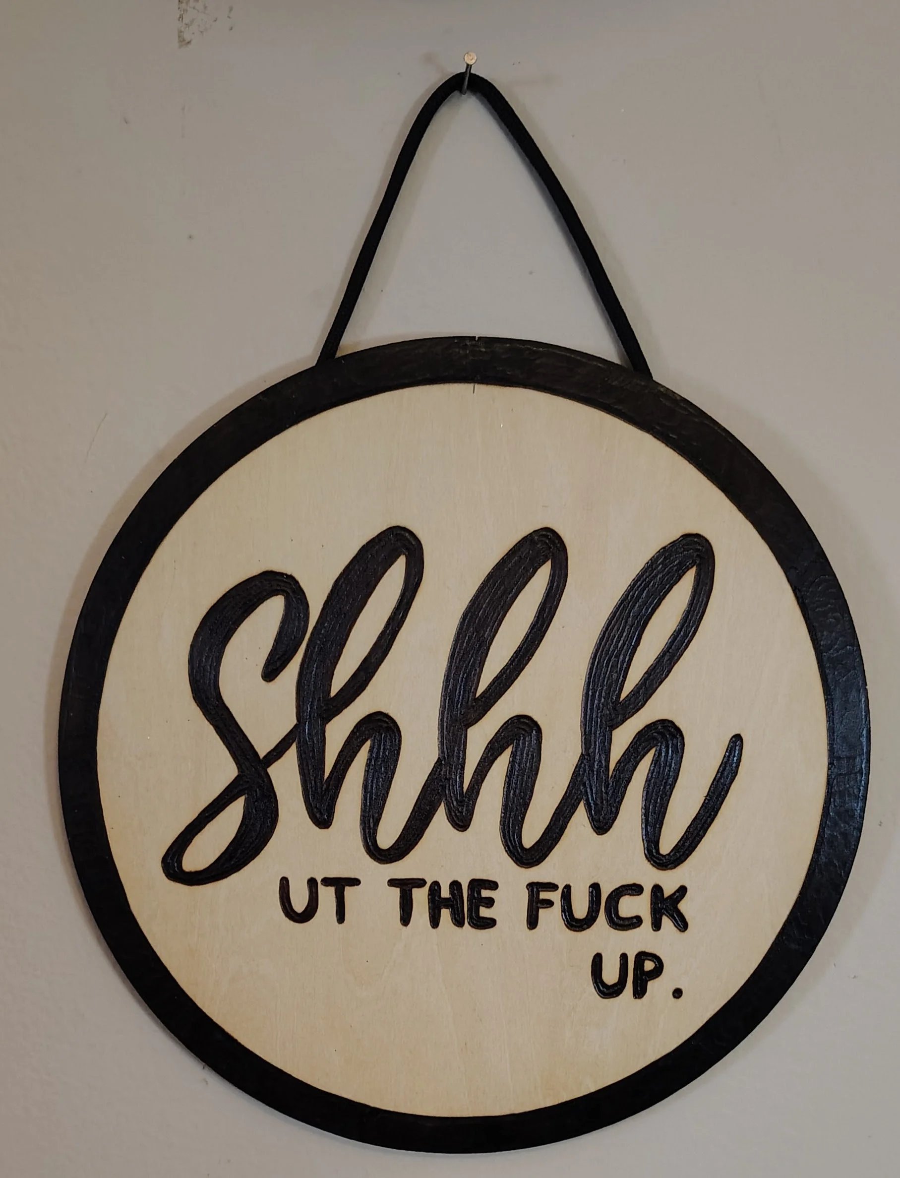 Circular wood burnt sign with black border and black loop hanger, inscribed with the words "shhh" and "UT THE F**K UP" in black lettering.